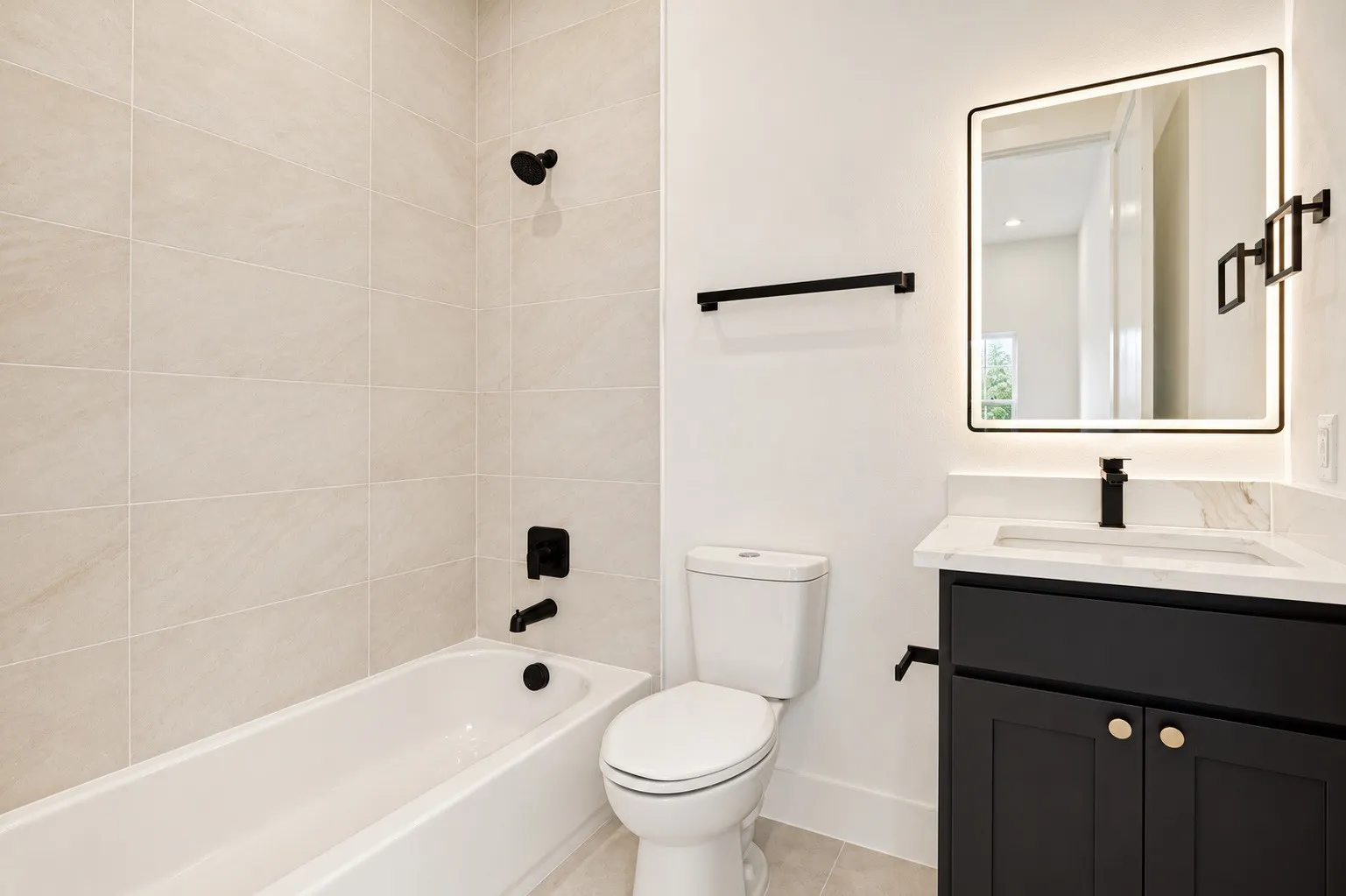 Soaking tub with matte black fixtures, neutral-toned tile surround, single vanity with black cabinetry, white countertop, and illuminated framed mirror