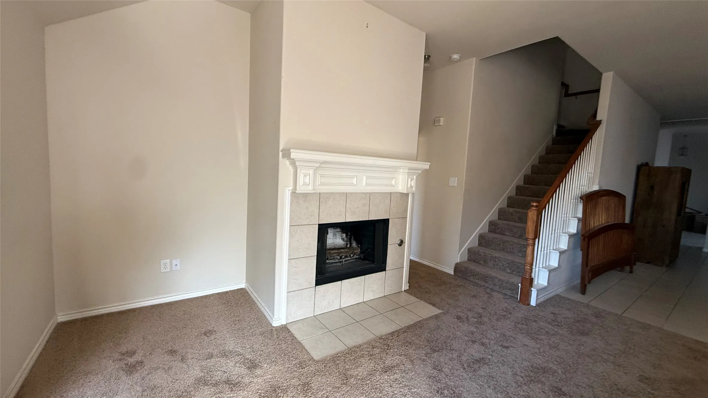 Tiled fireplace with a decorative mantel, carpeted flooring, and a staircase with a wood handrail