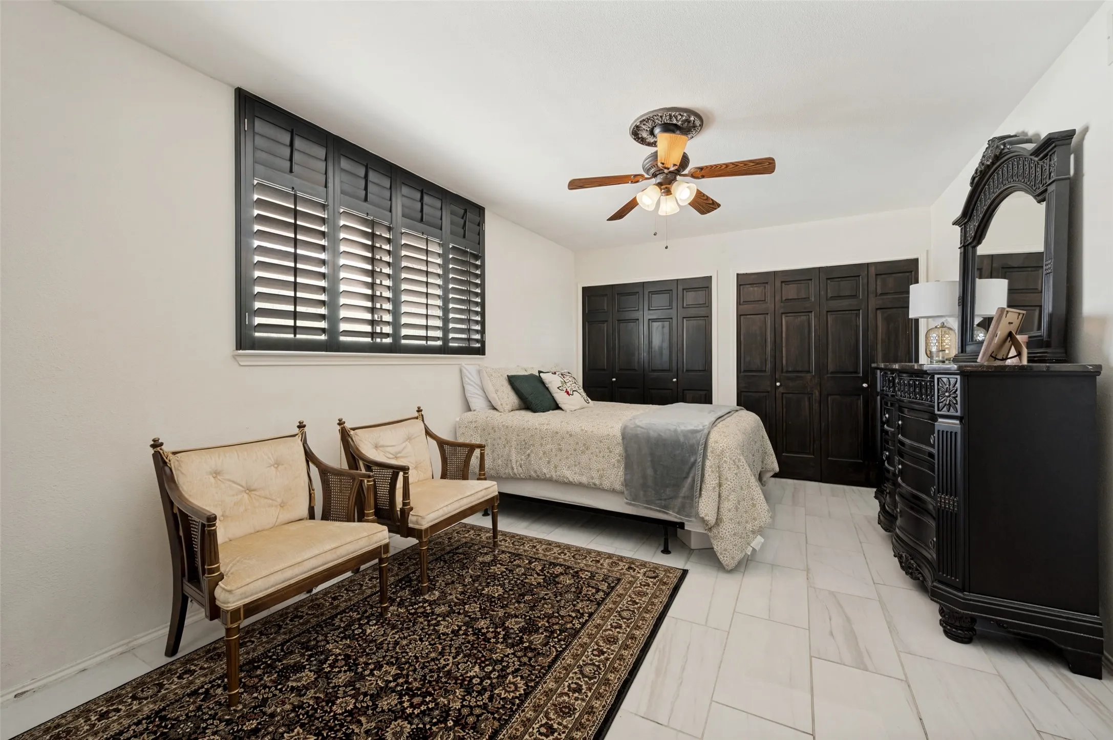 White tile flooring, black plantation shutters, two dark wood-finish bi-fold closet doors, and a ceiling fan with light fixture
