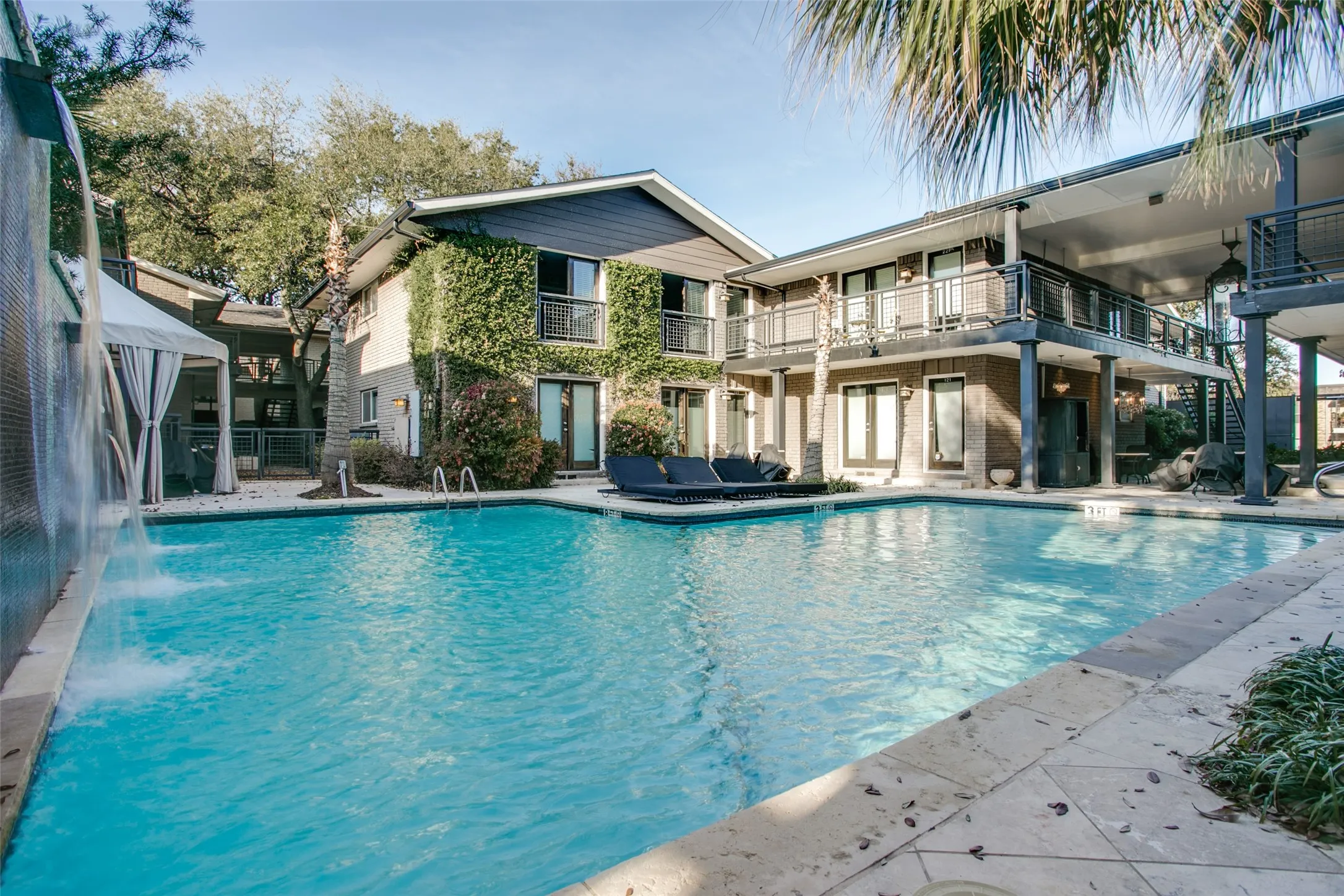 Swimming pool with a waterfall feature, surrounding architectural elements with balconies, and mature landscaping