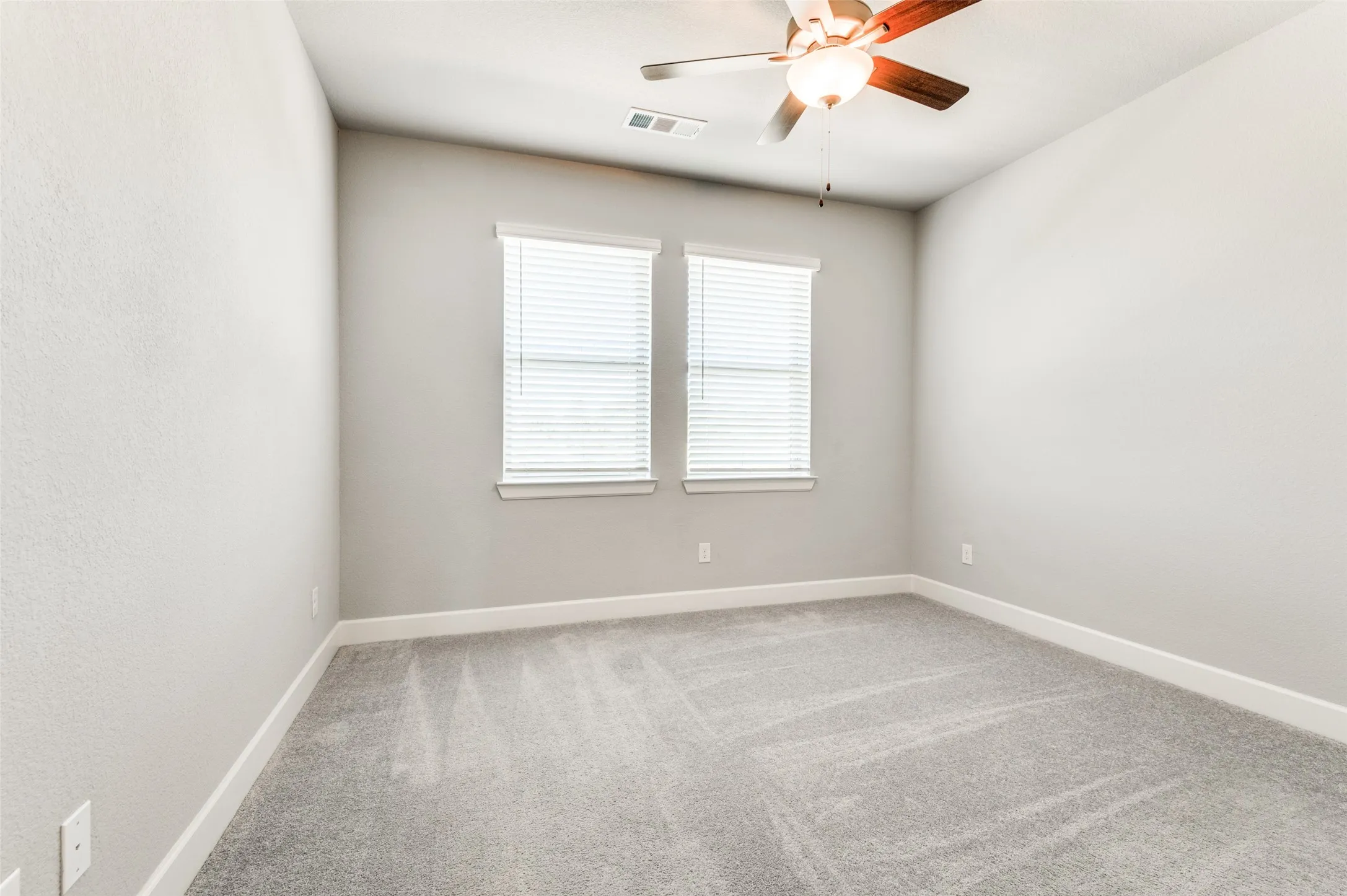 Neutral wall paint, two windows with blinds, ceiling fan with integrated lighting, and gray carpet flooring
