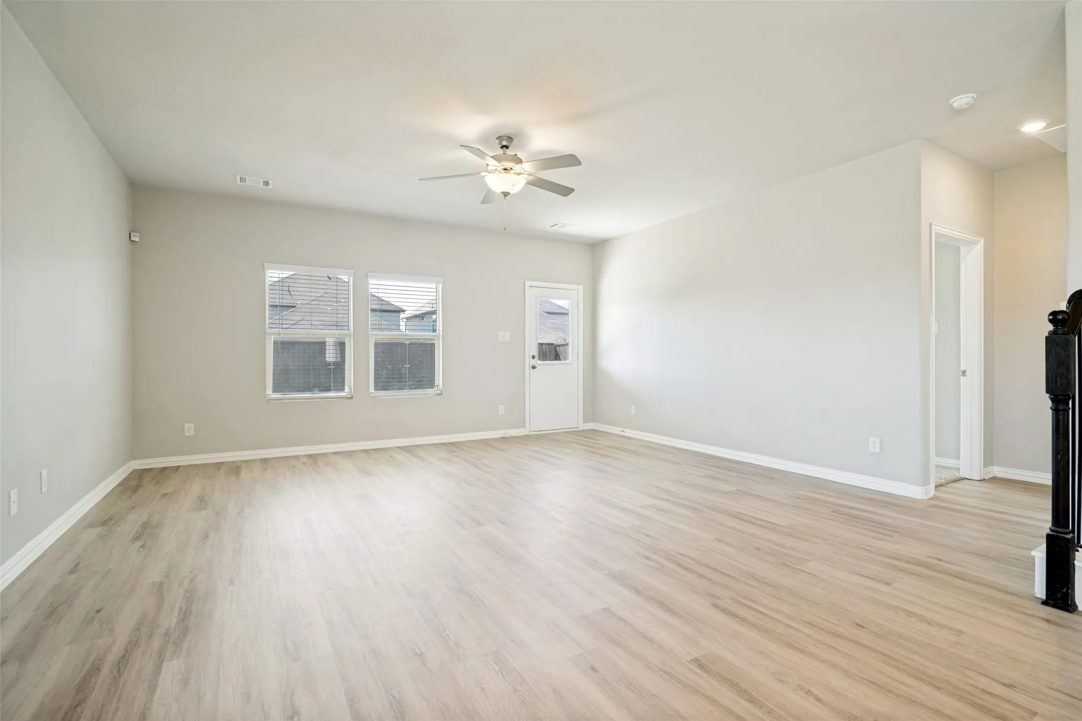 Spacious room featuring light-toned wood-finish flooring, multiple windows, a ceiling fan with integrated lighting, and a white door with an exterior light