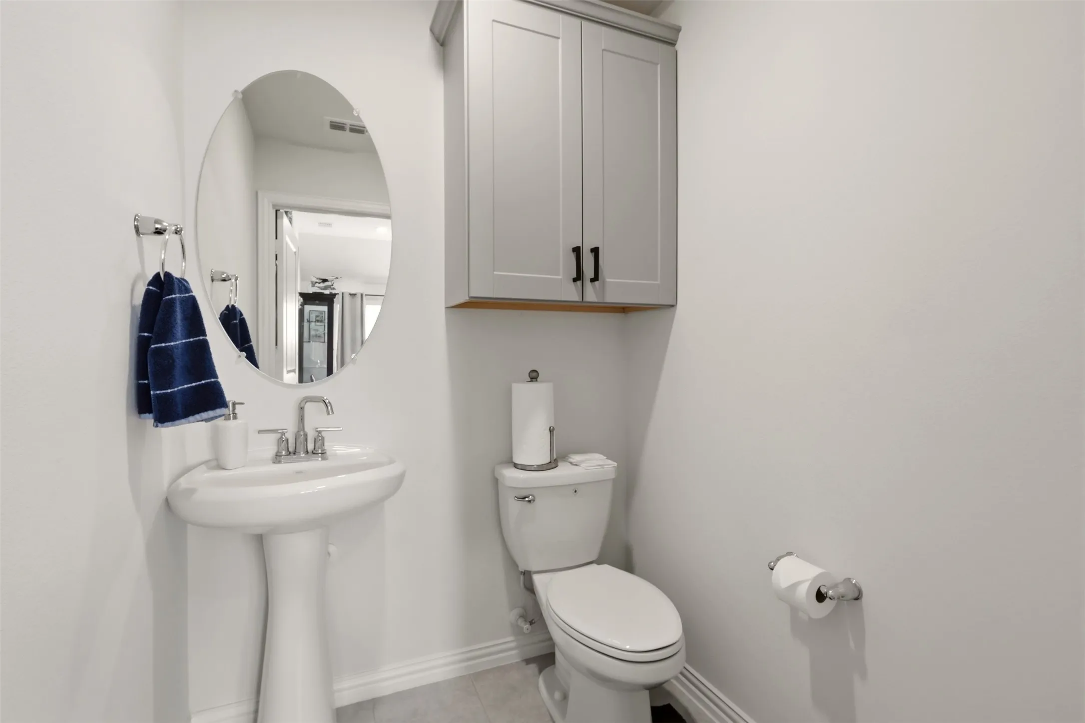 Pedestal sink with chrome faucet, oval wall-mounted mirror, and wall-mounted cabinetry with matte black hardware