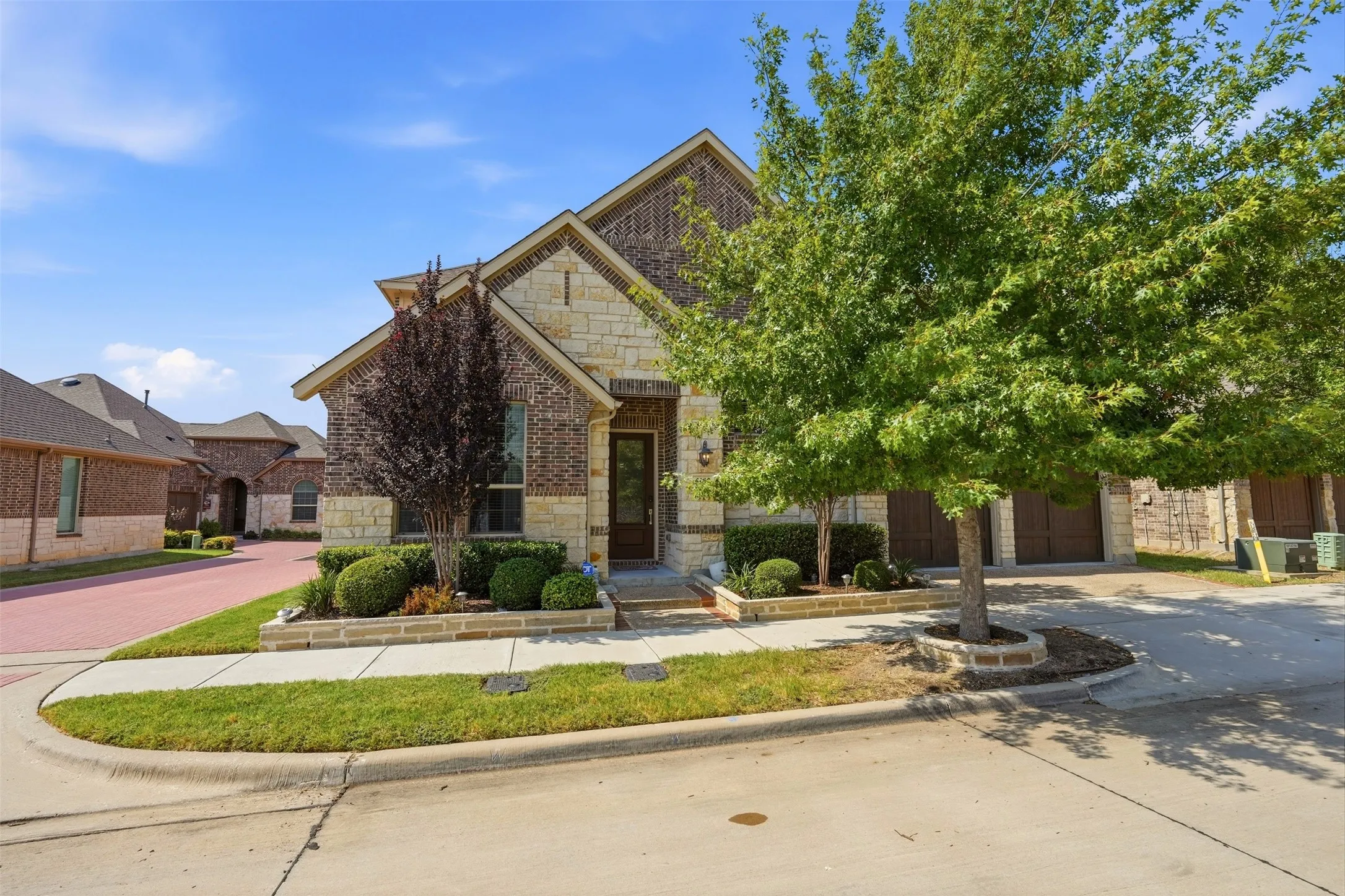 Brick and stone facade, gabled roofing, integrated planting beds, paved driveway, and established trees