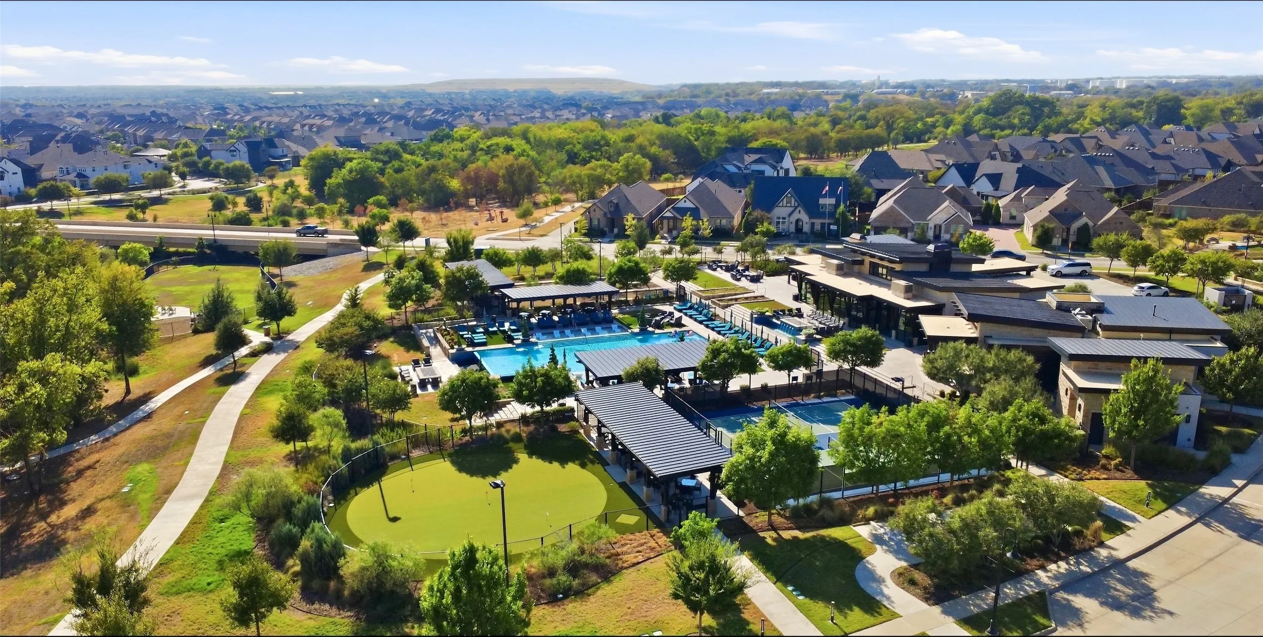 Resort-style amenities featuring a large outdoor swimming pool, tennis court, putting green, and expansive community building with additional covered outdoor spaces, all surrounded by residential properties and mature landscaping