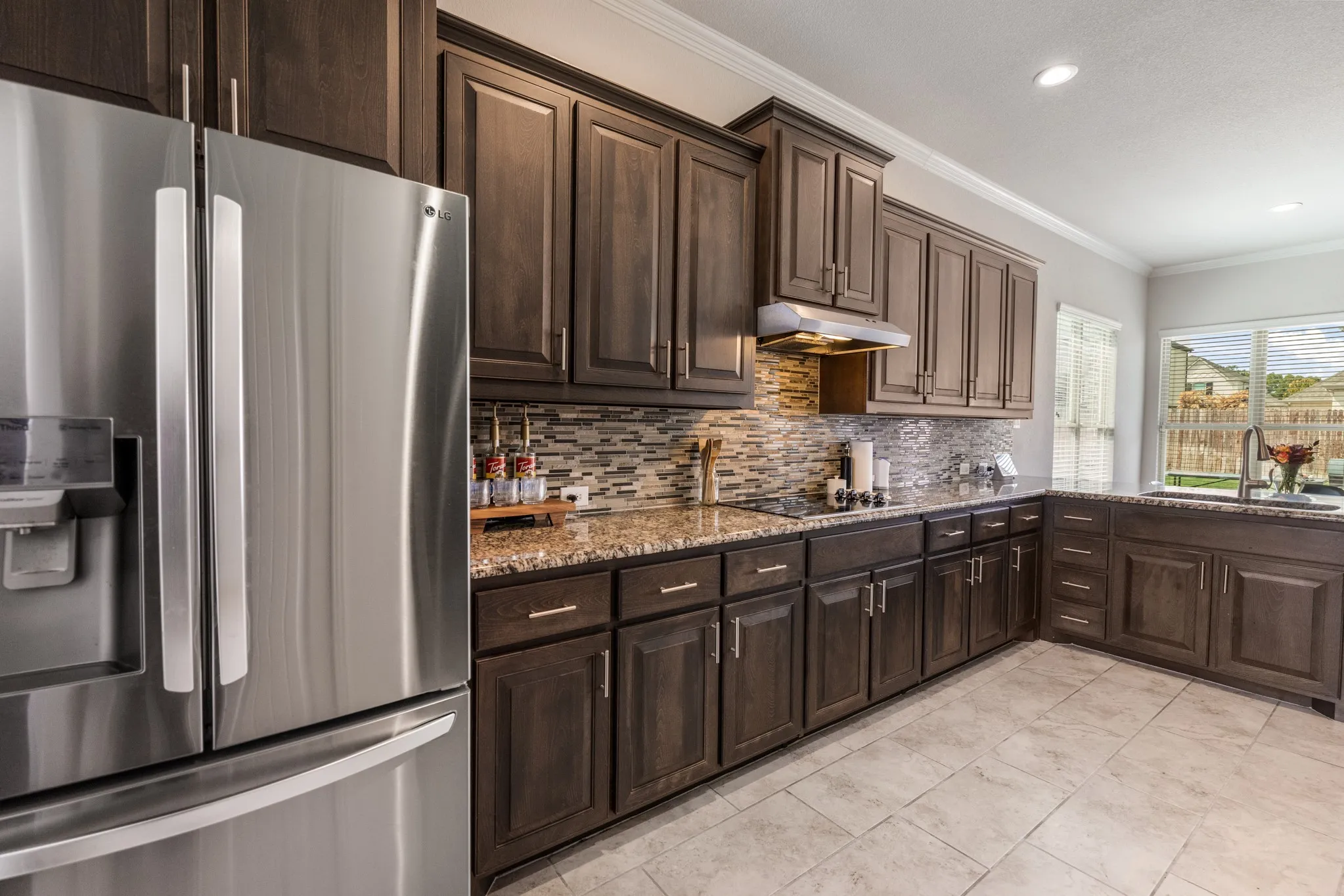 Stainless steel refrigerator, dark wood cabinetry, patterned tile backsplash, light-colored tile flooring, and a large window above the sink