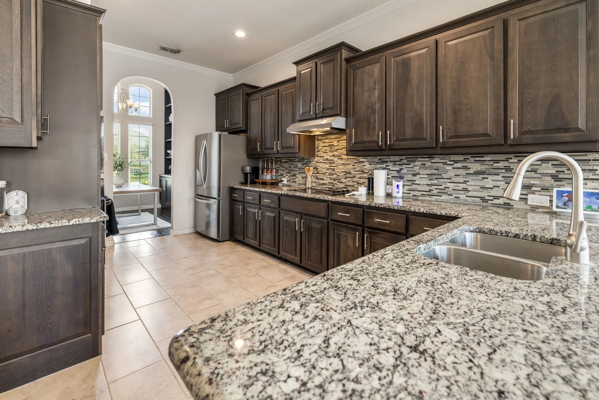 Kitchen featuring extensive dark wood cabinetry, granite countertops, stainless steel appliances, a double basin sink with a high-arch faucet, and a mosaic tile backsplash