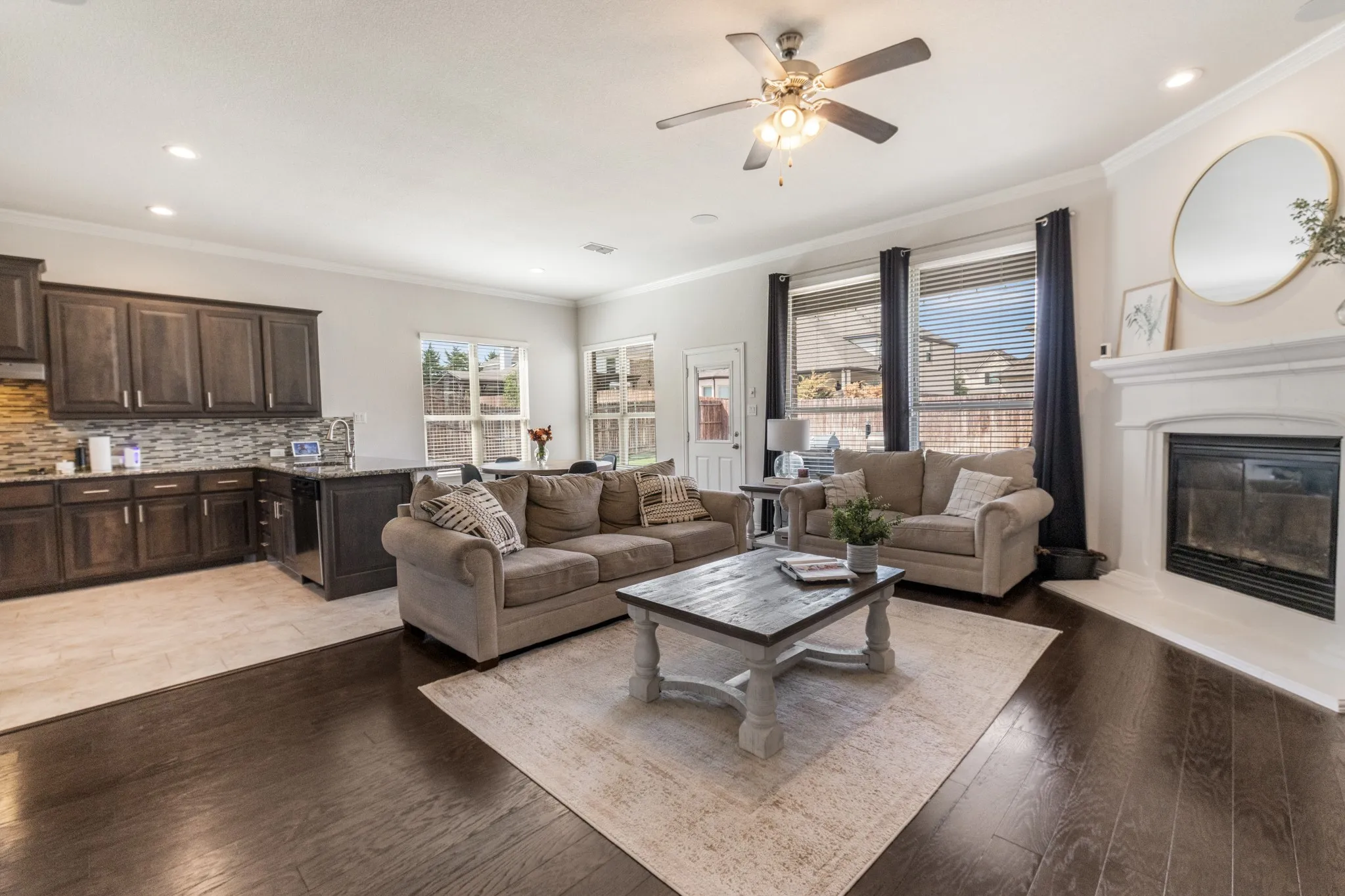 Open-concept living area featuring rich wood-finish flooring, a contemporary ceiling fan with integrated lighting, and a fireplace with a white mantel