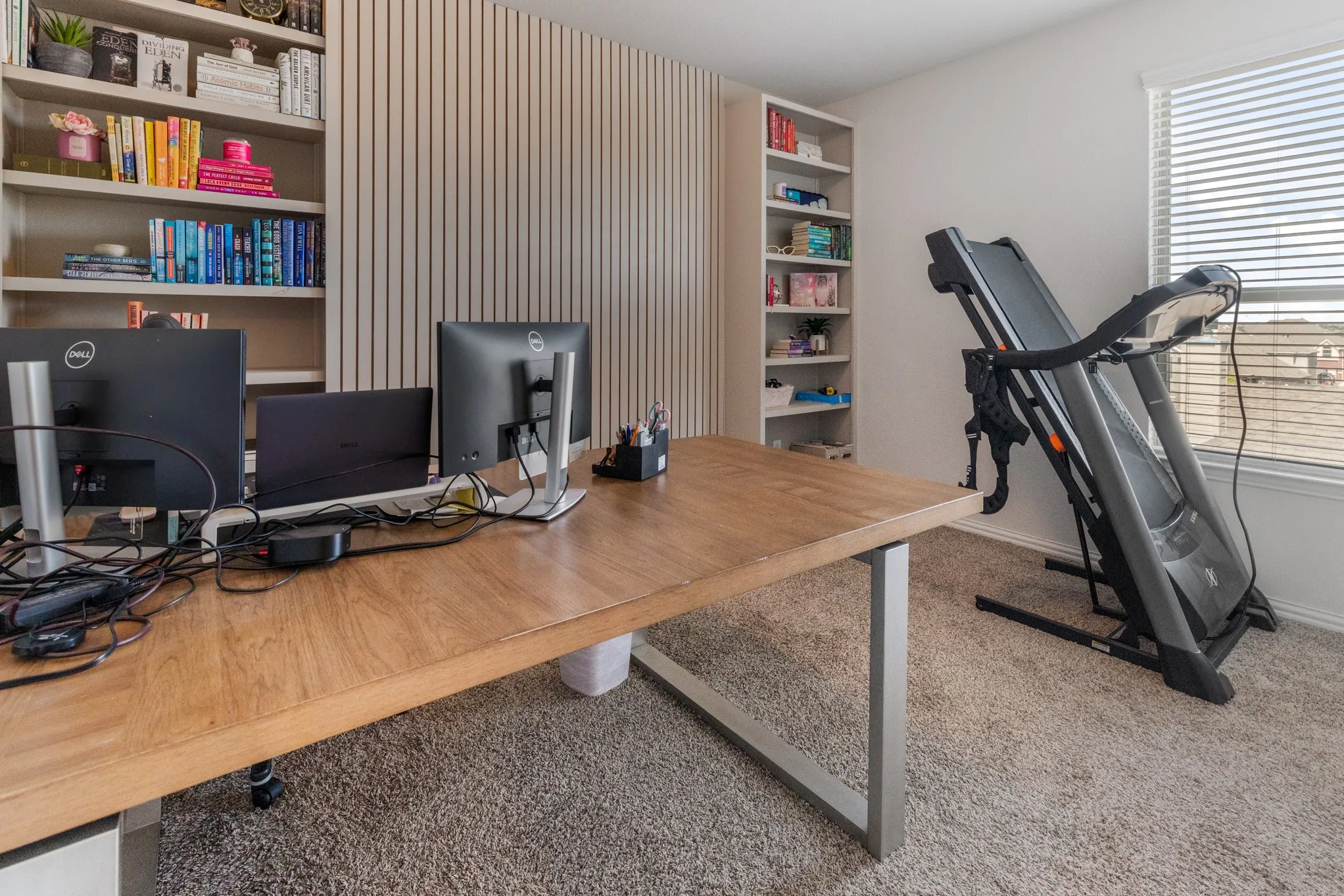 Modern home office featuring a spacious wood-finish desk, built-in shelving with vertical slat wall paneling, dual monitor setup with ergonomic workspace, natural light from large windows with blinds, and neutral carpeting throughout