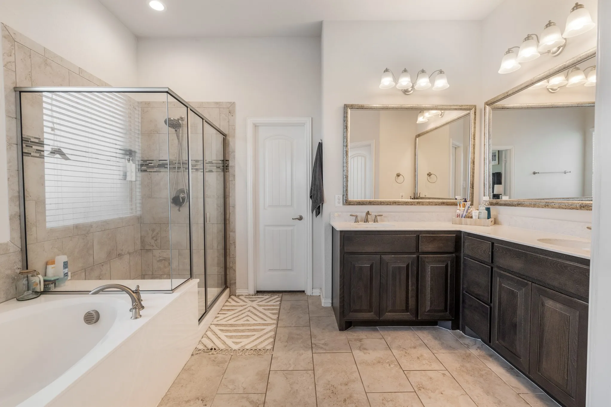 Tiled flooring throughout, dual wood-finish vanity with countertop, framed mirrors, glass-enclosed shower, and built-in soaking tub