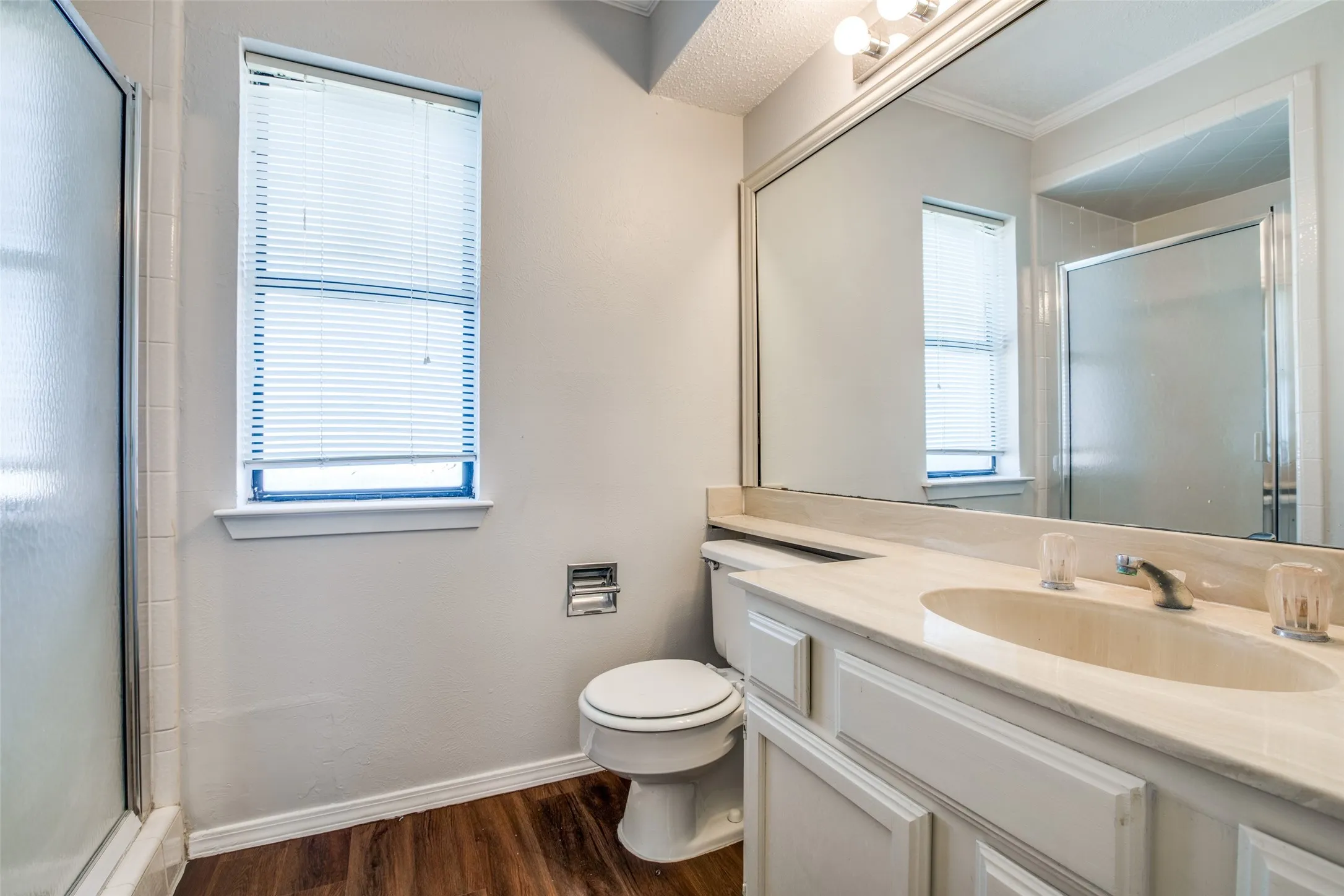 Bathroom featuring wood-finish flooring, a vanity with an integrated sink, a large wall-mounted mirror, a window with blinds, and a shower with a glass enclosure