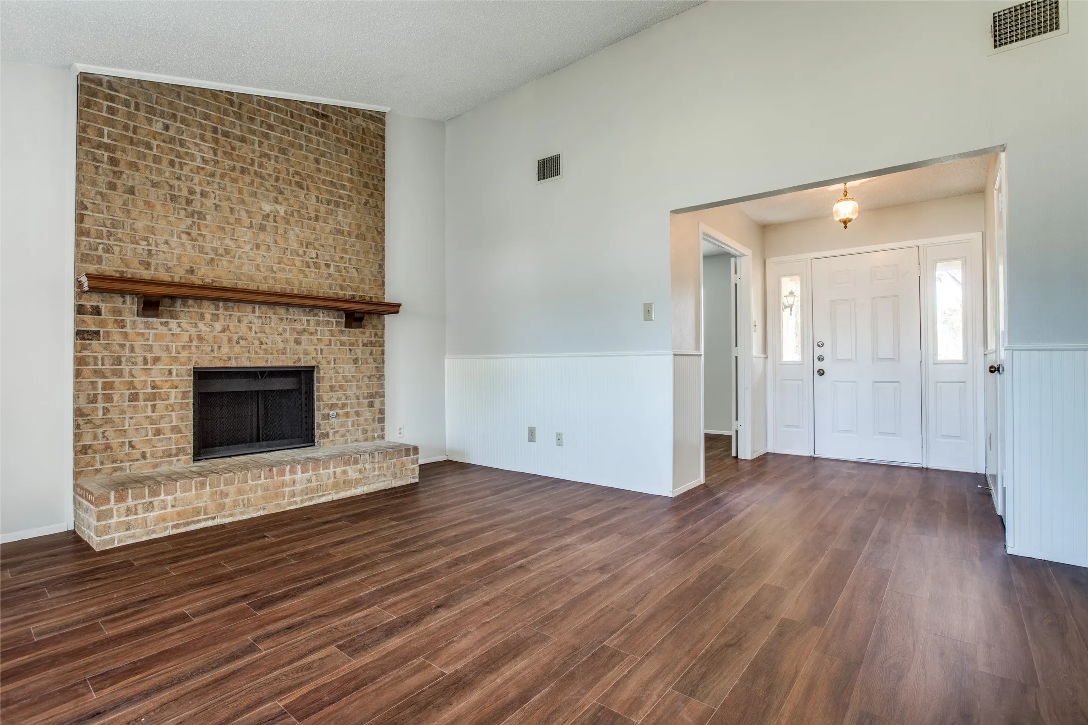 Expansive living area with wood-finish flooring, a brick fireplace with a wood mantle, and wainscoting