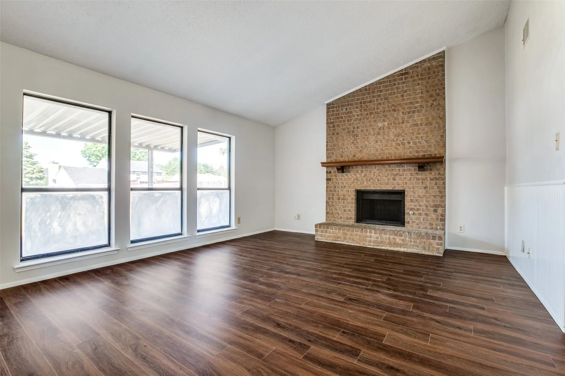 Brick fireplace with a wood-finish mantle, dark wood-finish flooring, and multiple large windows