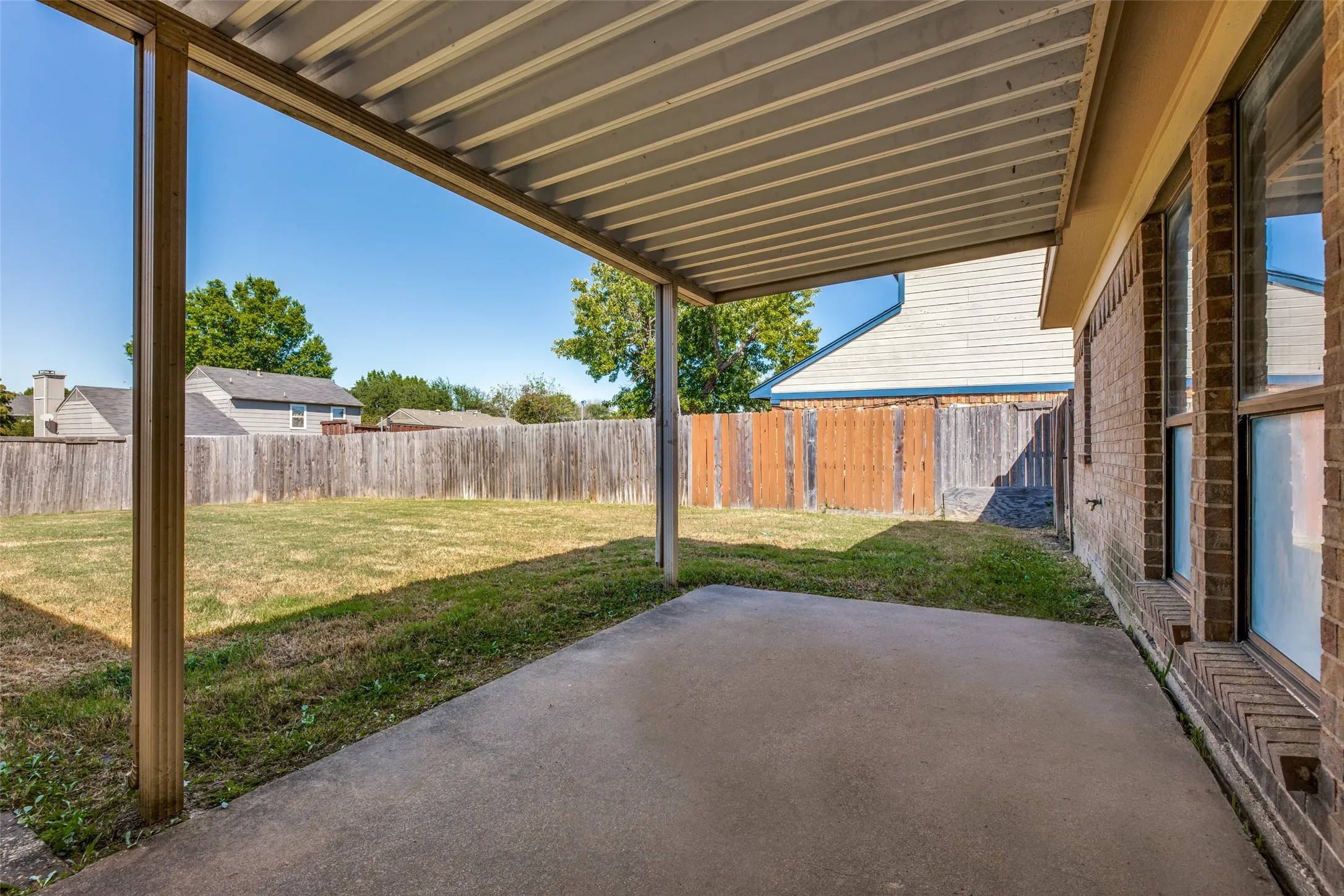 Covered concrete patio with a metal awning, offering direct access to a fenced backyard featuring a grassy area