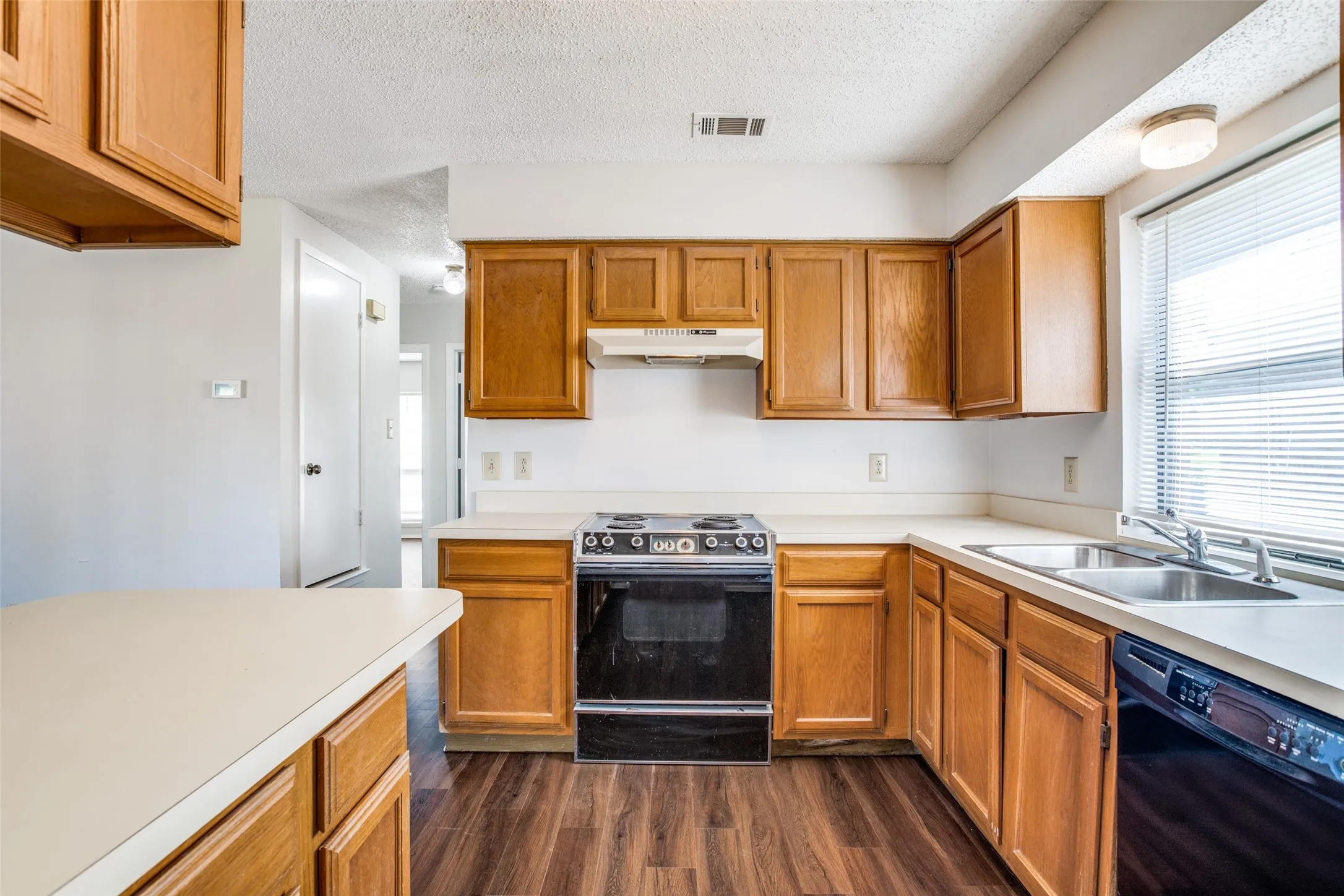 Kitchen featuring wood-finish cabinetry, light-toned countertops, a stainless steel double basin sink, and wood-finish flooring