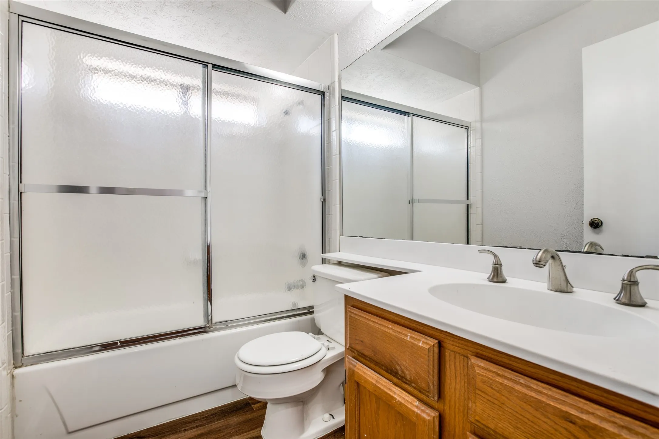 Wood-finish flooring, full vanity with integrated sink, brushed nickel faucet, large wall mirror, and a tub/shower combination with sliding frosted glass doors
