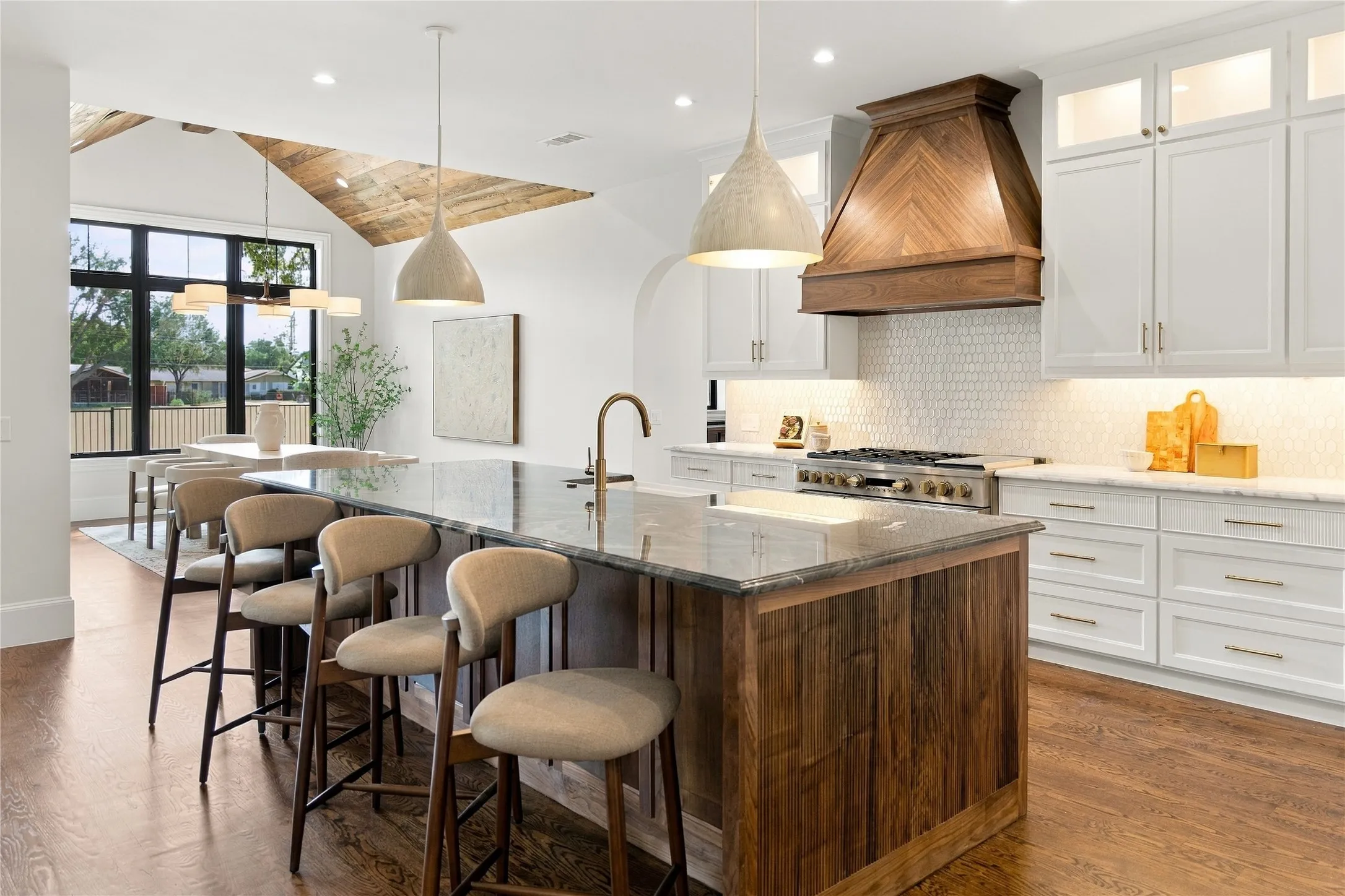 Spacious kitchen featuring a large island with a stone countertop, white cabinetry with gold-tone hardware, a wood-finish range hood, and wood-finish flooring