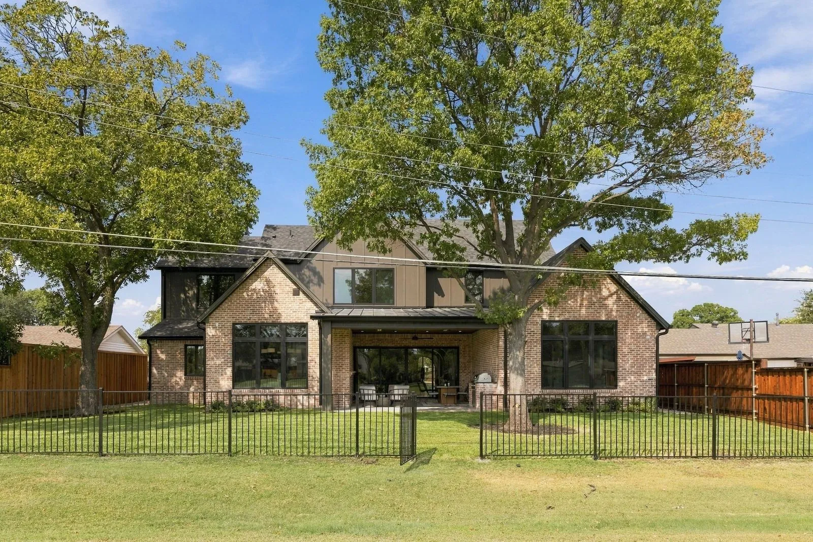 Brick and siding exterior with a covered patio, expansive lawn, mature trees, and black metal fencing