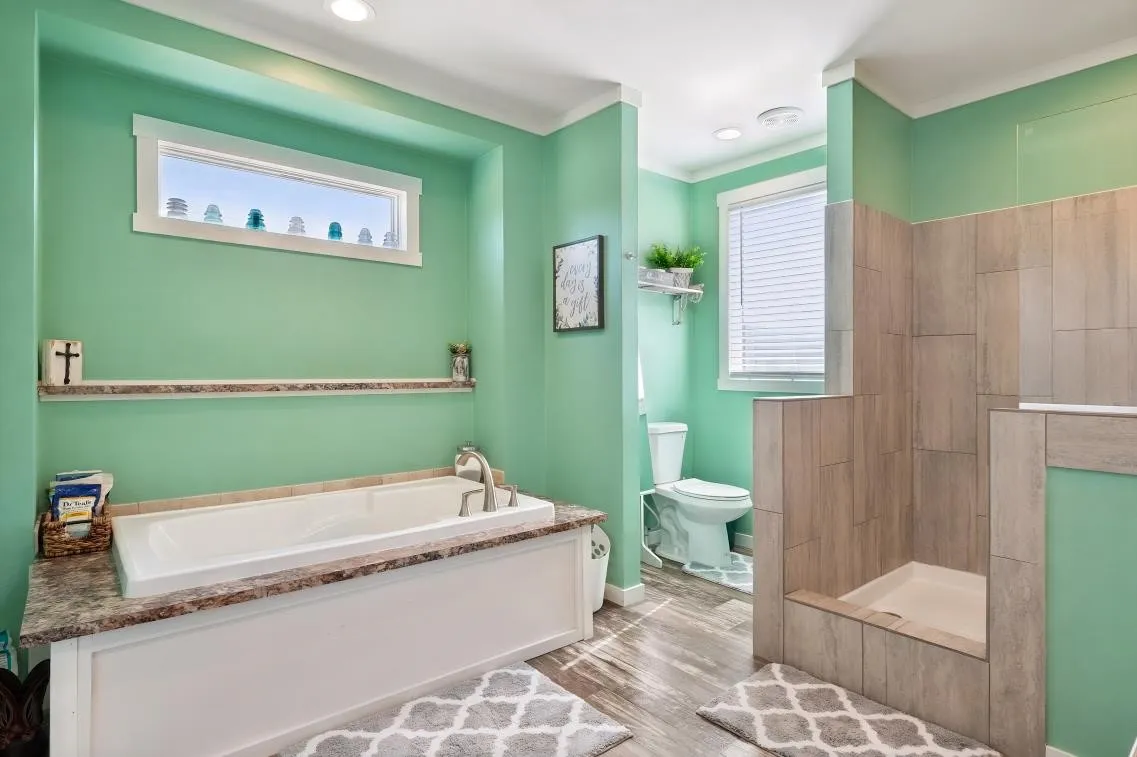 Built-in soaking tub with a stone-finish surround, walk-in shower with tile surround, wood-finish flooring, and a horizontal window