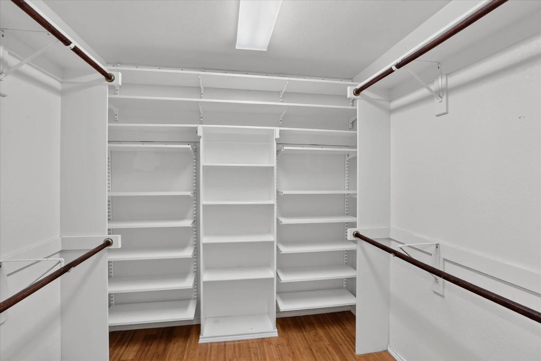 Walk-in closet featuring wood-finish flooring, white built-in shelving, white walls, and clothes hanging rods