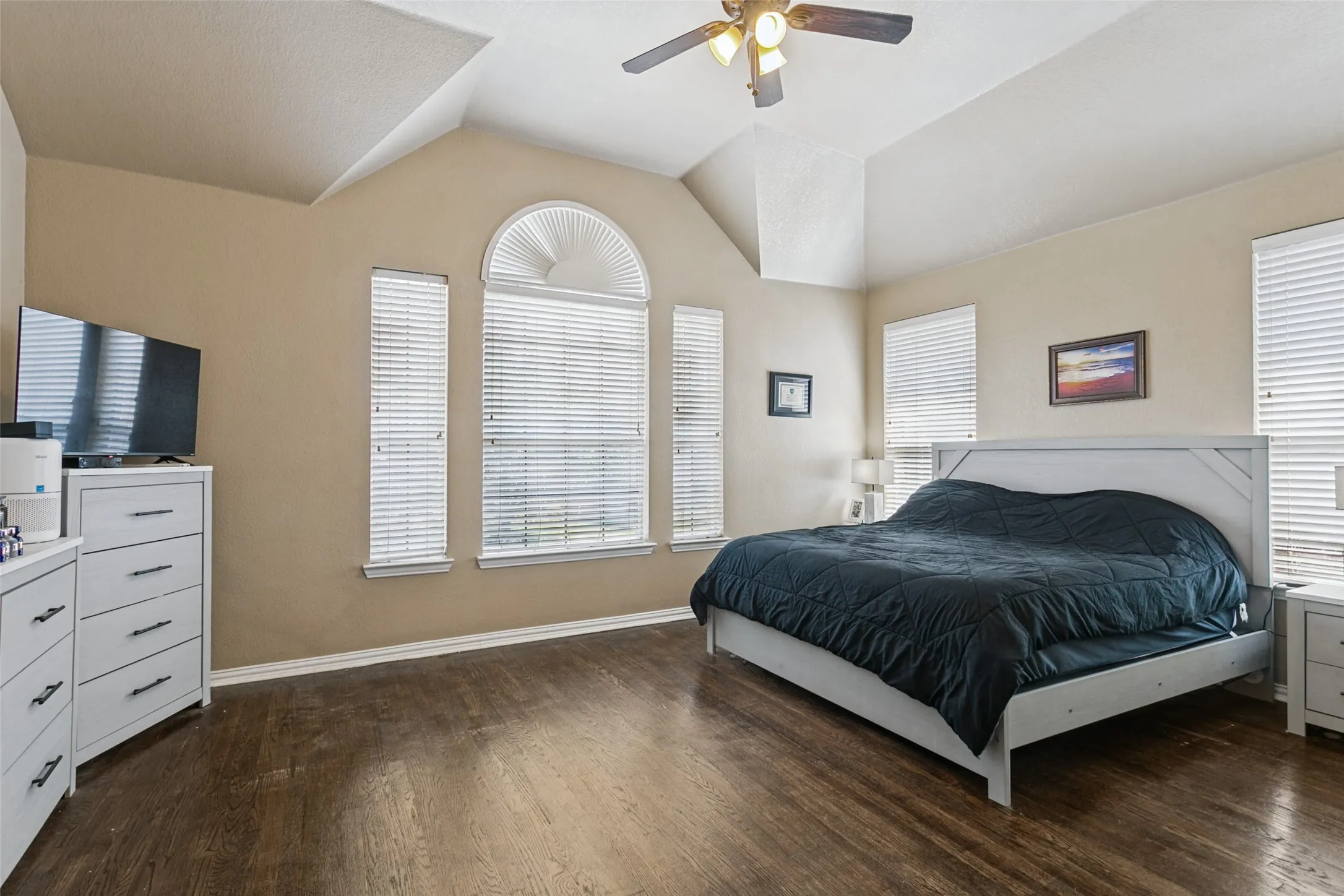 Vaulted ceiling with a ceiling fan, hardwood-finish flooring, multiple windows with blinds, and an arched window