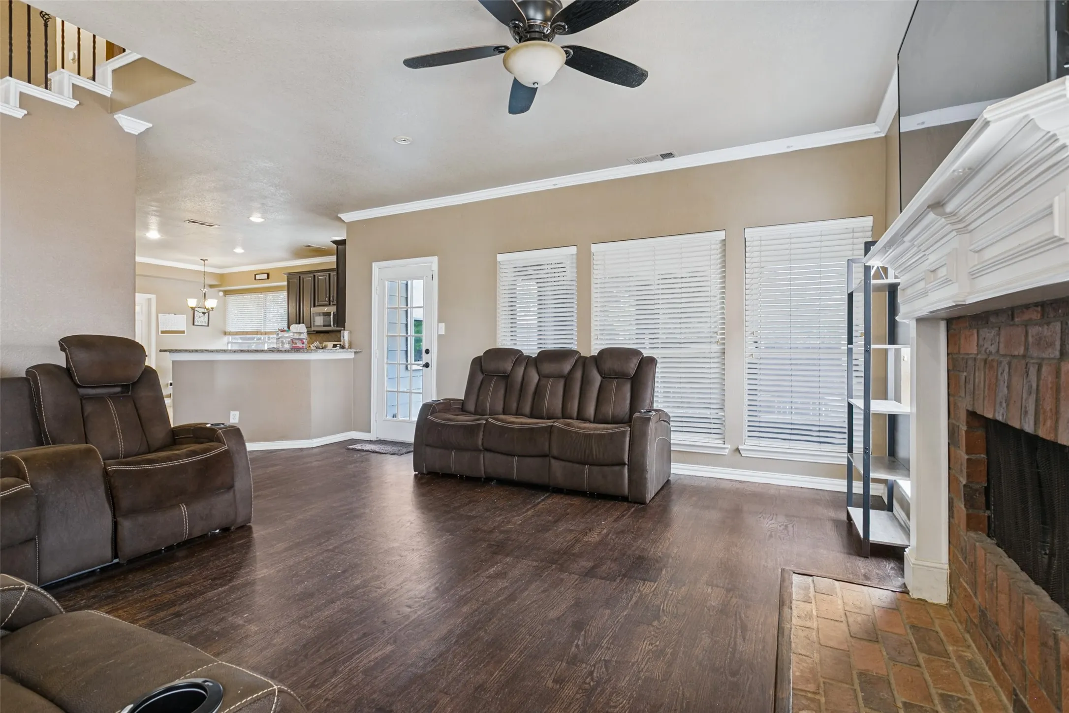 Wood-finish flooring, brick fireplace with a white mantel, ceiling fan with integrated lighting, multiple windows with blinds, and white trim molding