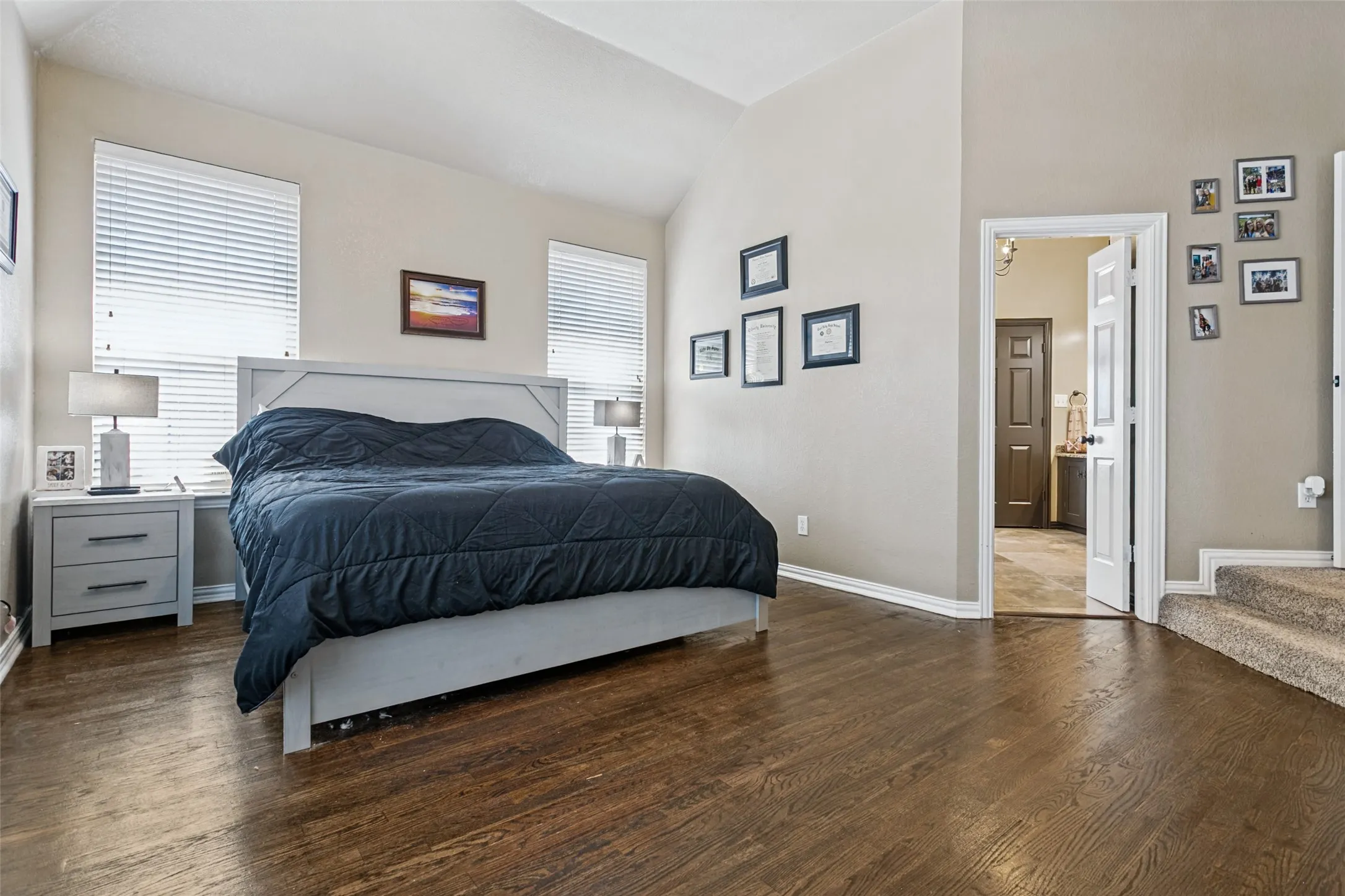 Hardwood-style flooring, vaulted ceiling, two windows with blinds, open doorway, and carpeted stairs