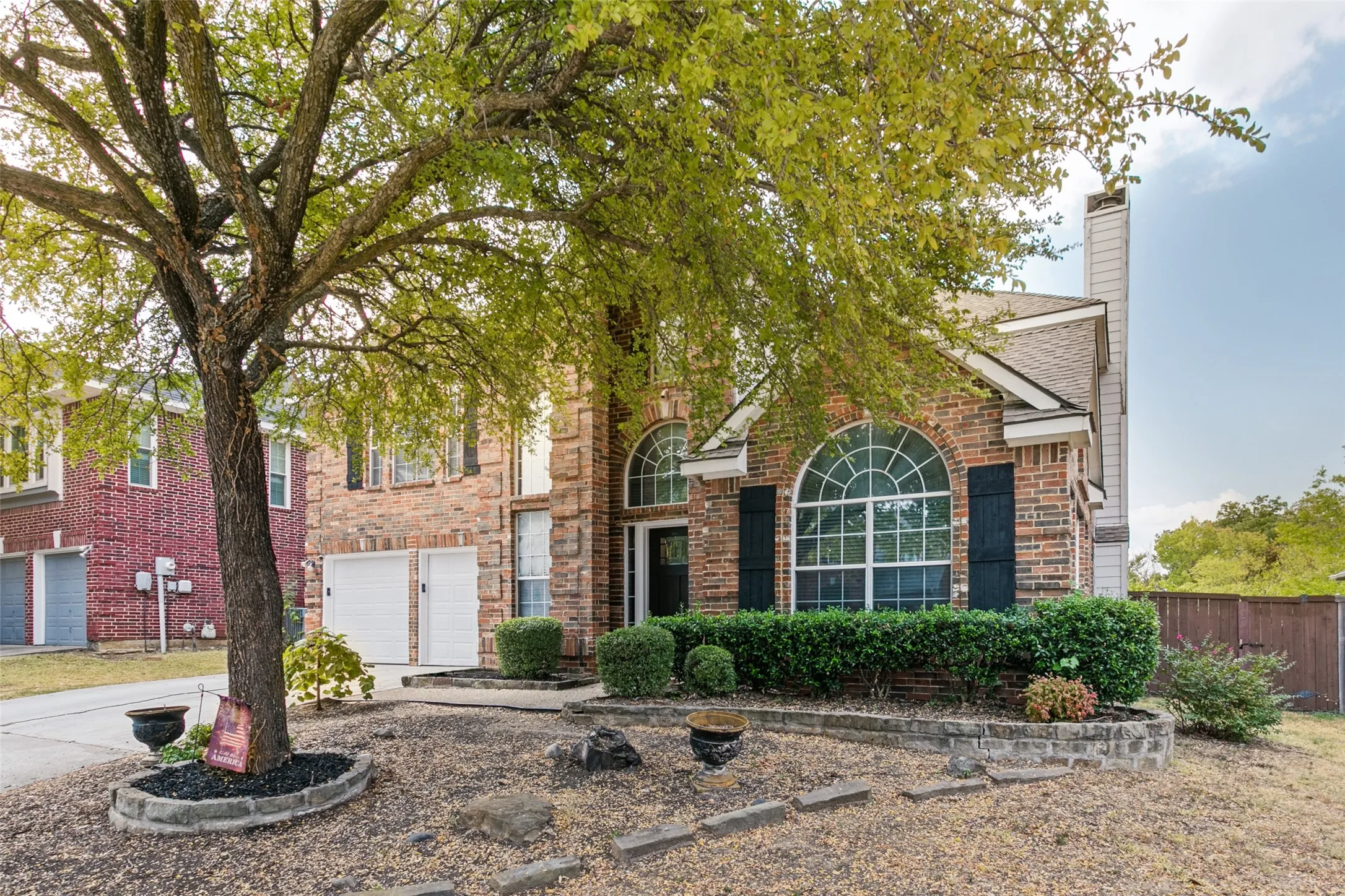 Brick and stone façade with arched windows and shutters, two-car garage, mature tree, and landscaped front yard