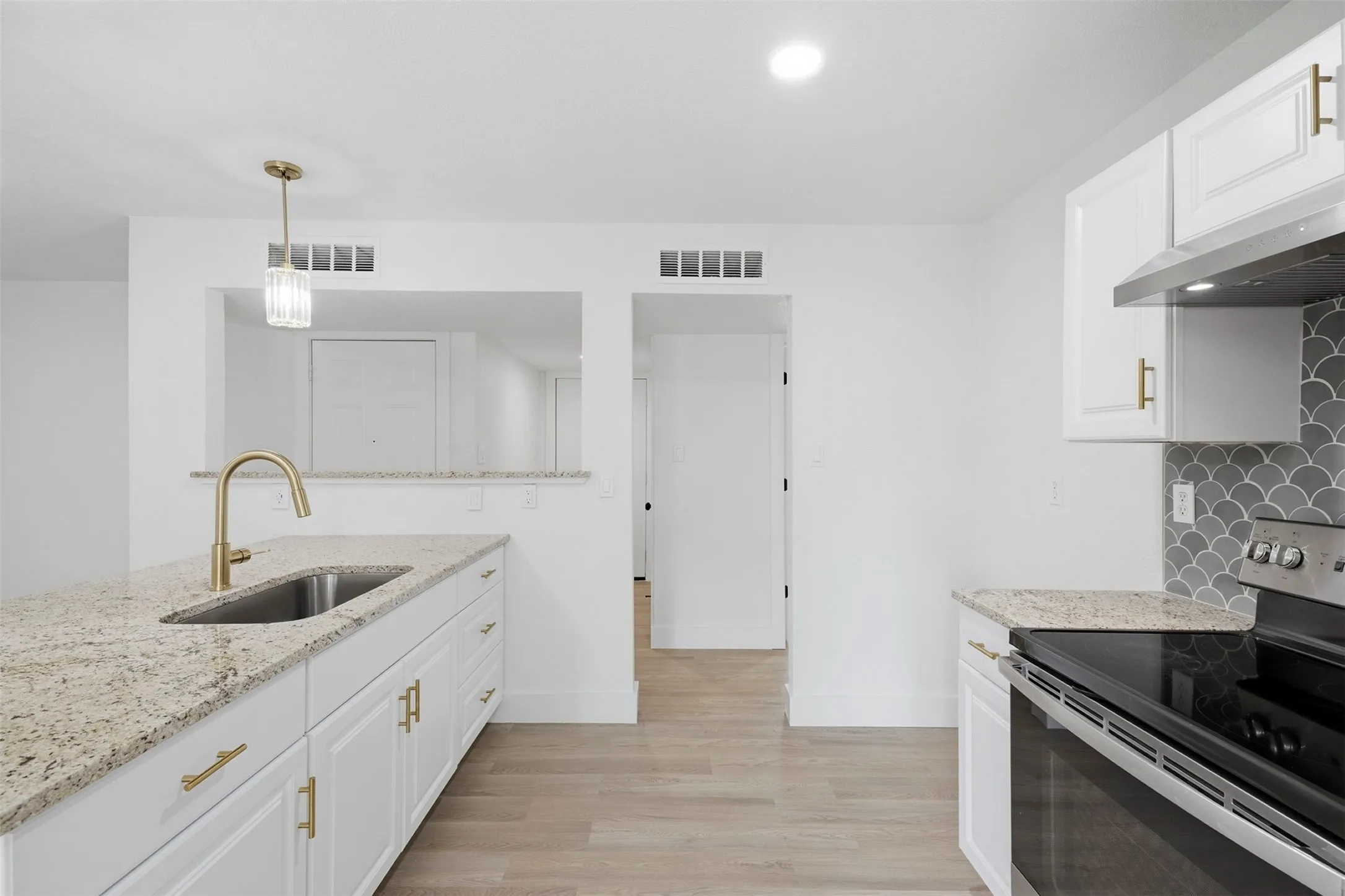 Kitchen featuring granite countertops, white cabinetry with gold-tone hardware, wood-finish flooring, a stainless steel sink with a gold-tone faucet, and a scallop-patterned backsplash