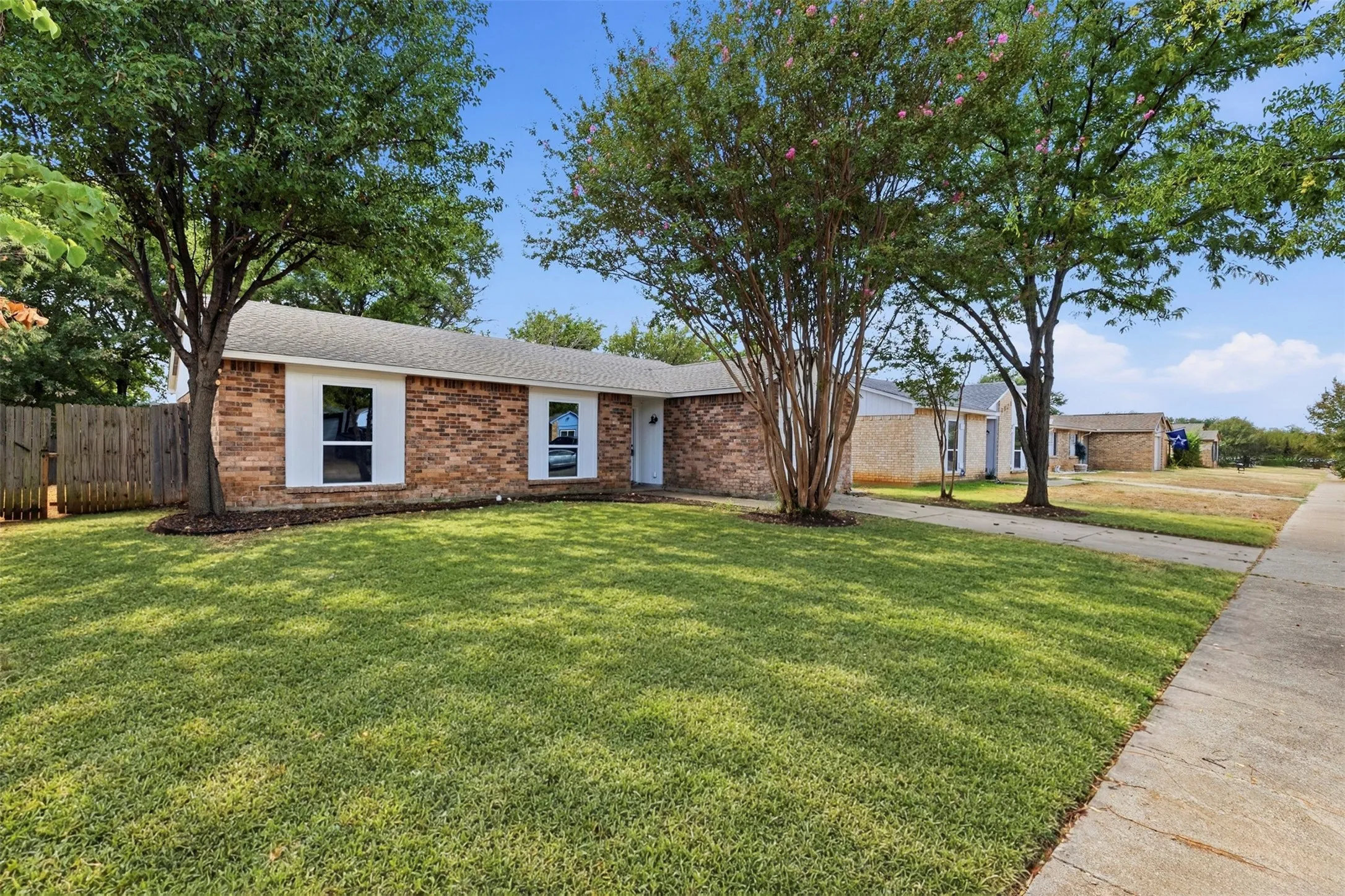 Brick facade, white window shutters, white front door, mature trees, and a concrete walkway