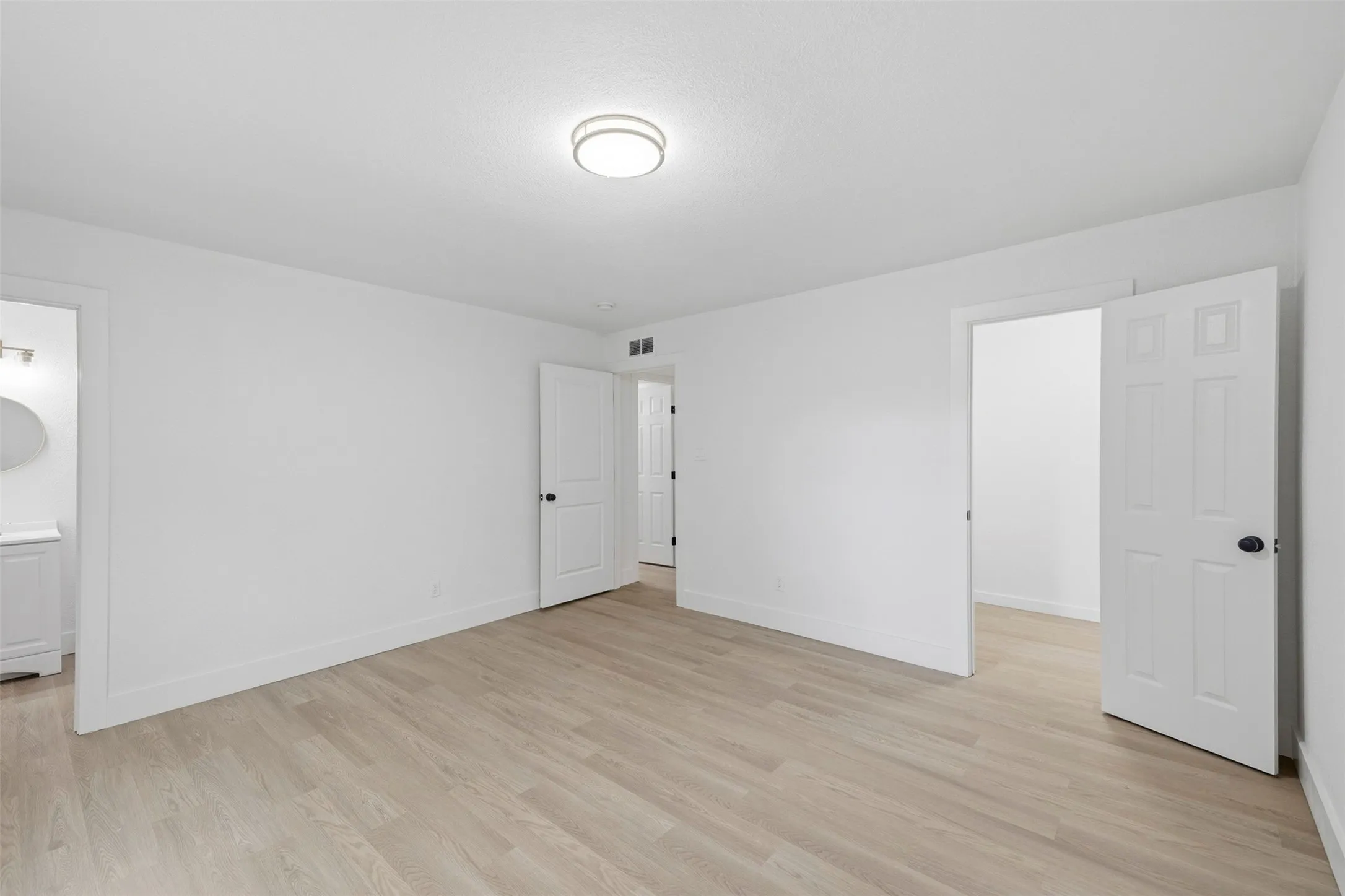 Recessed ceiling light fixture, light wood-finish flooring, white walls, white interior doors with black hardware, and an adjacent bathroom with a vanity and wall-mounted mirror