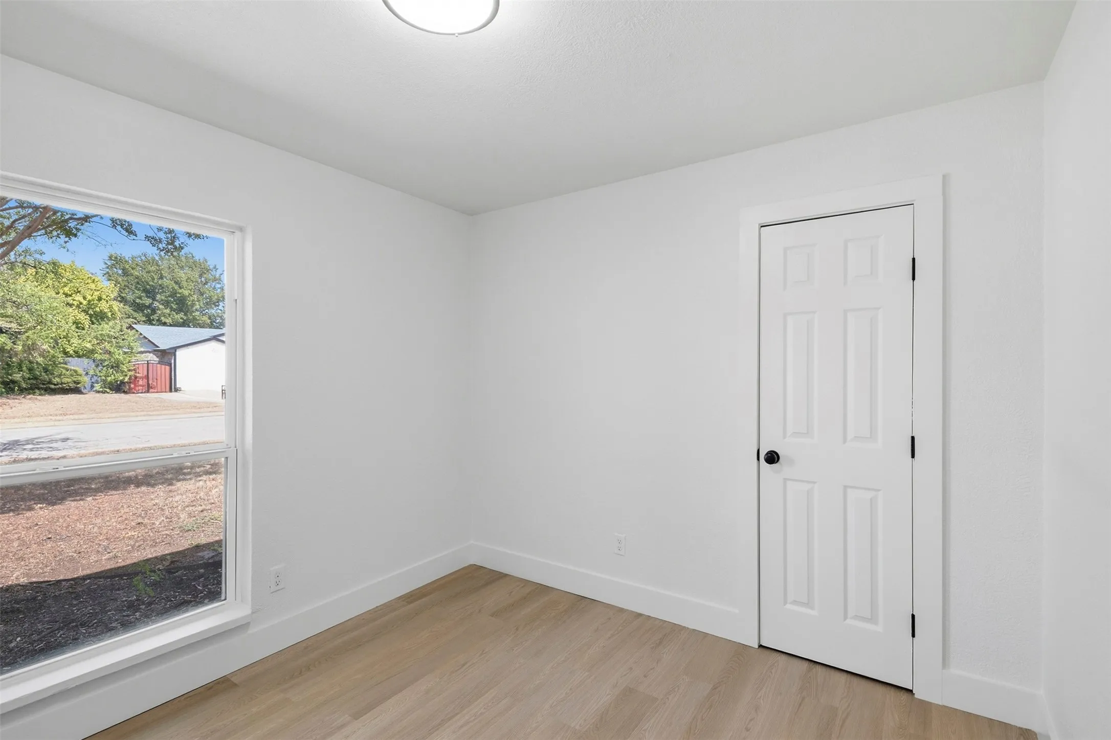 Wood-finish flooring with white baseboards, a white paneled door with a matte black knob, and a window with white trim