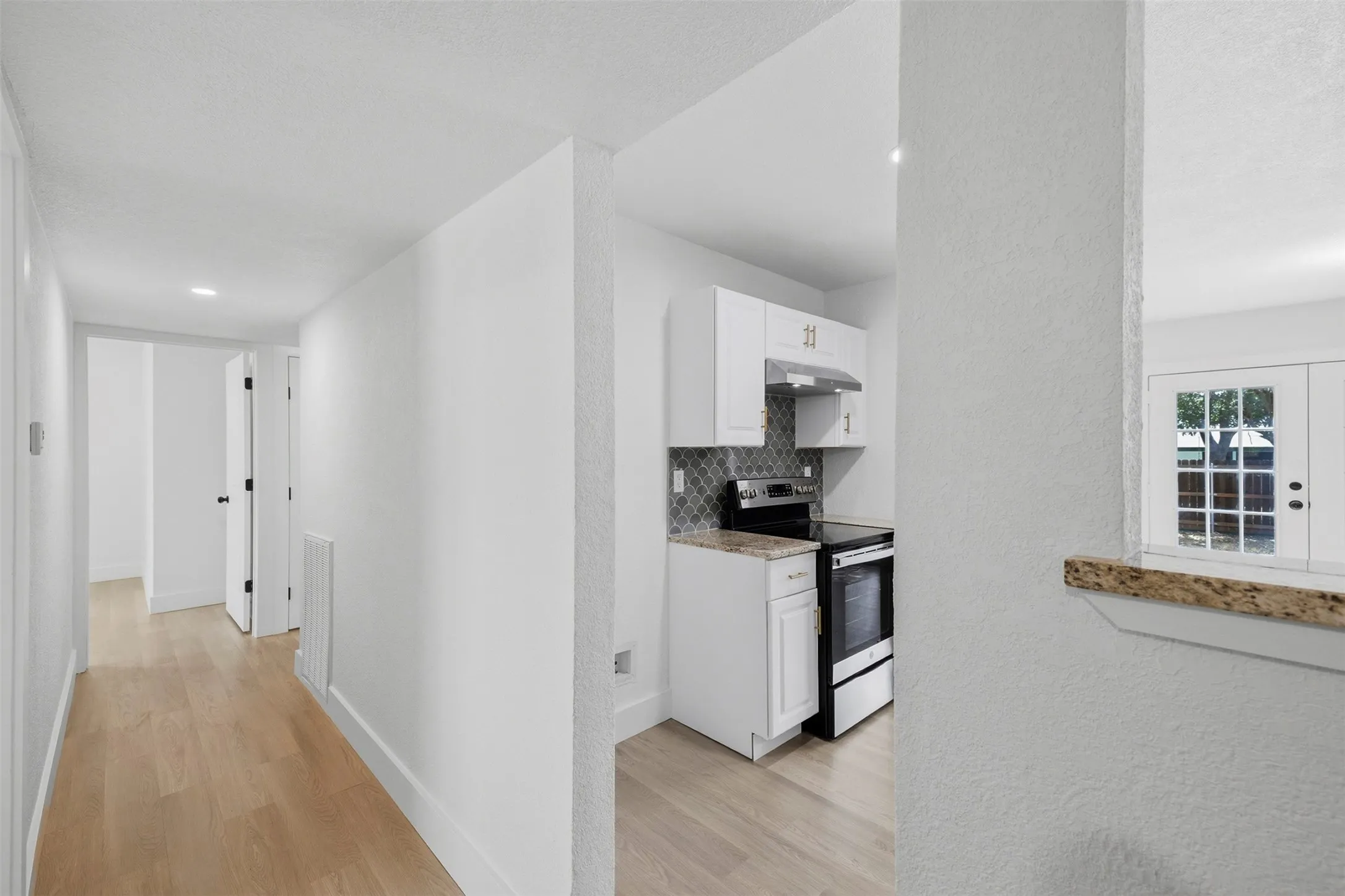 Wood-finish flooring, white walls, and a kitchen featuring white cabinetry, a tile backsplash, and a built-in oven with a range hood
