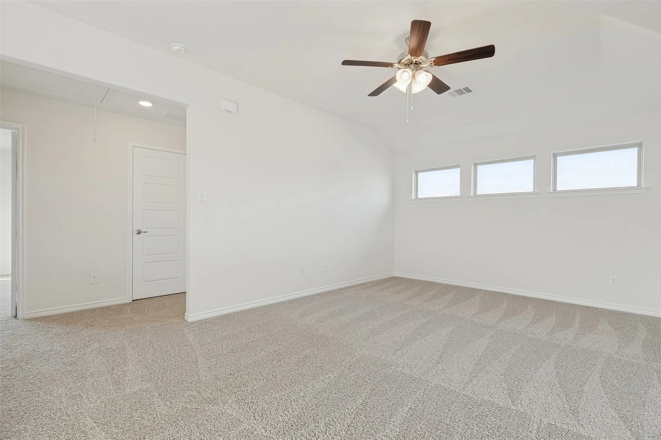 Expansive room featuring vaulted ceilings, light-tone carpet, a ceiling fan with light fixture, three high-set windows, and a panel door with brushed nickel hardware