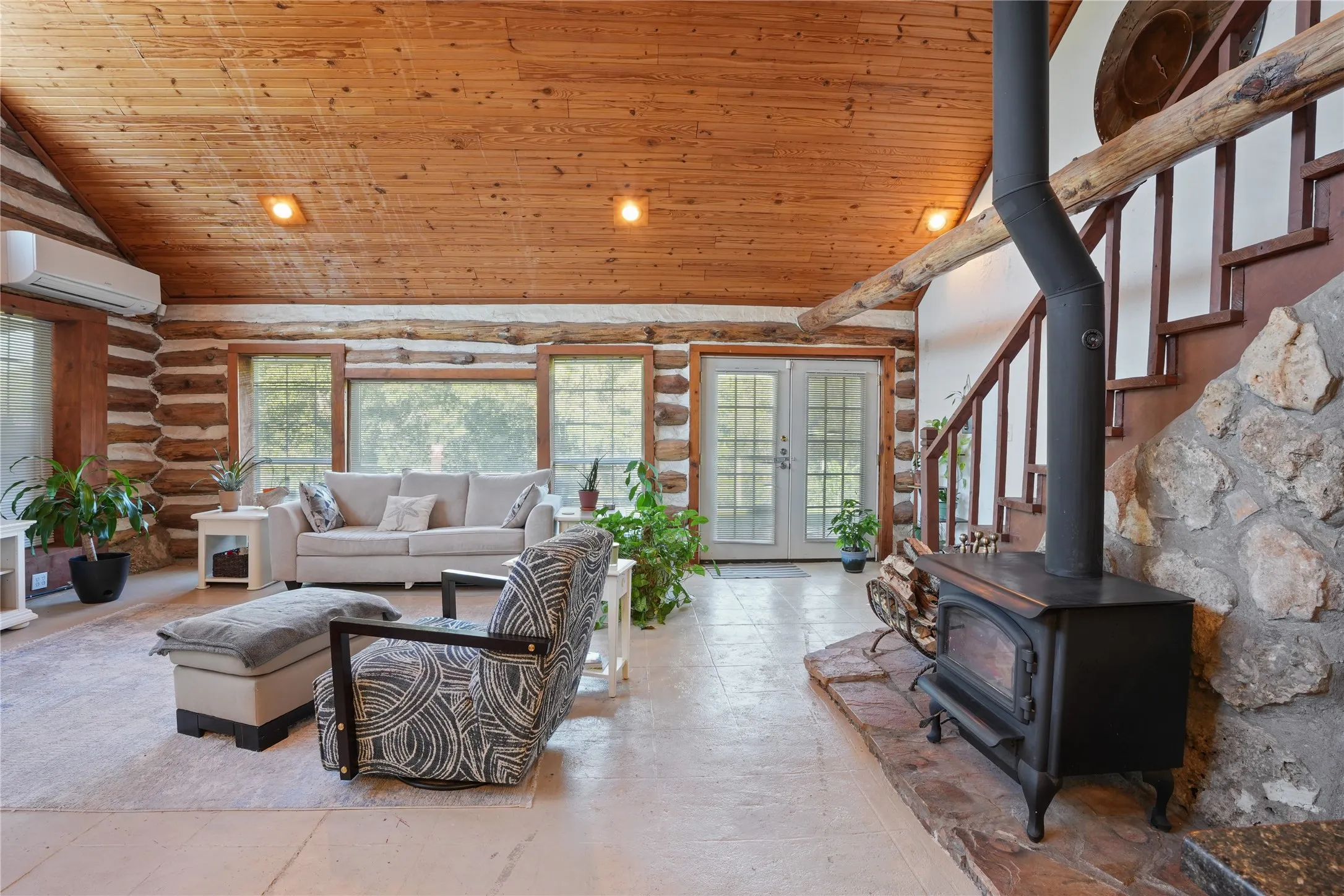 Wood-plank ceiling, exposed log walls, multiple windows, stone hearth, and wood-burning stove