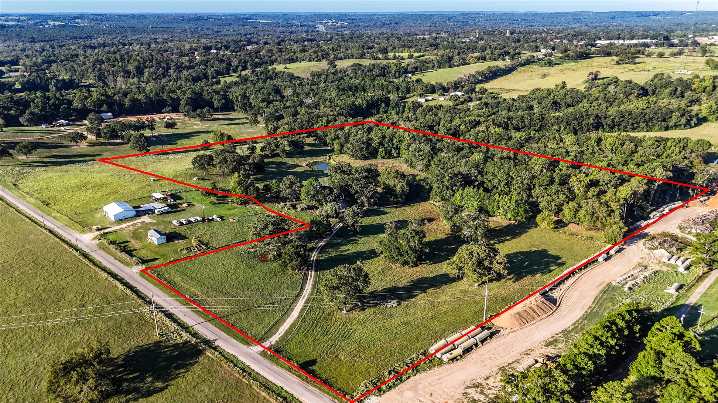 Expansive land featuring a mix of open fields and mature tree coverage, bordered by a paved road and a dirt road