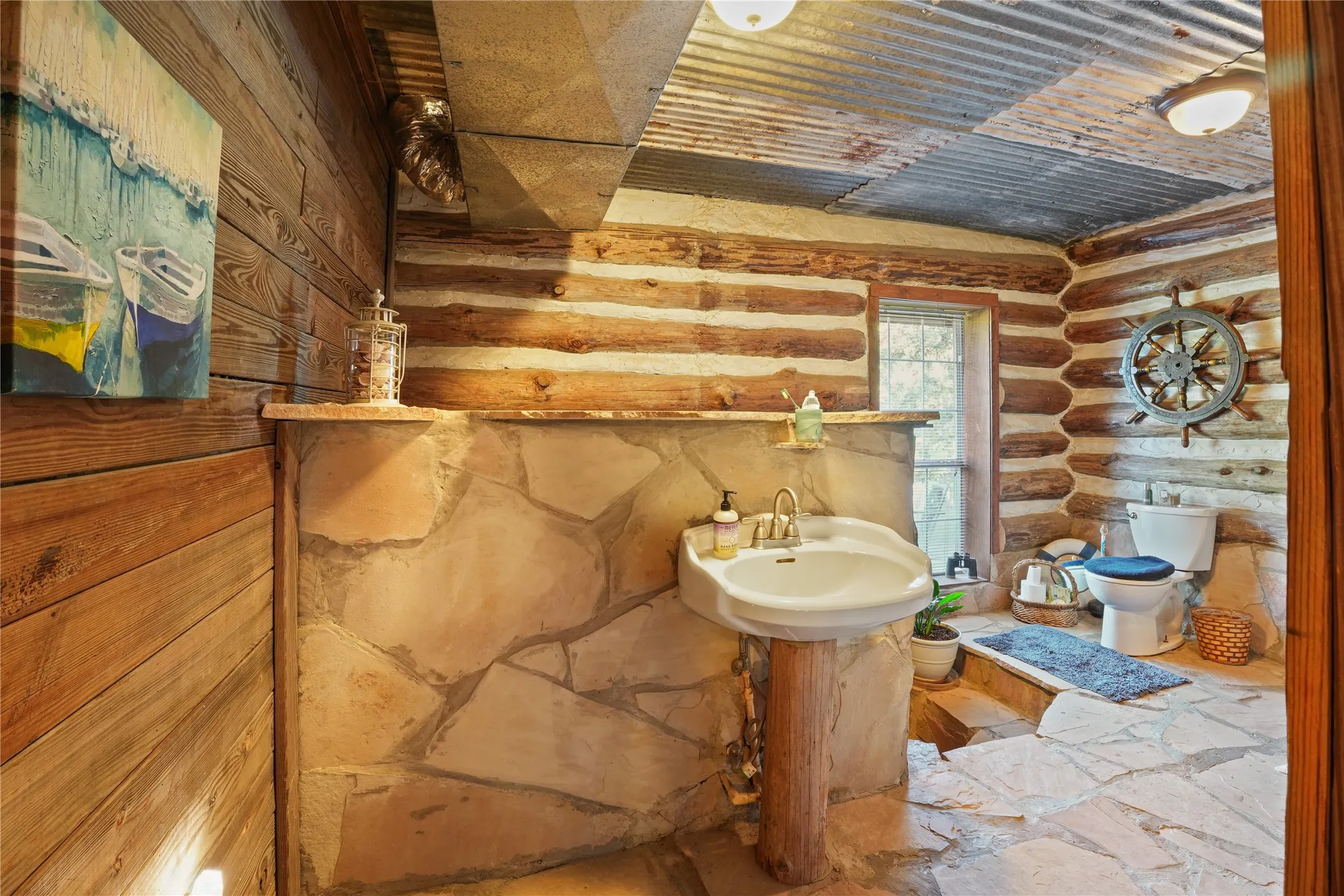 Log walls with chinking, stone-finish flooring, corrugated metal ceiling, pedestal sink with a wood base, and a clear glass window