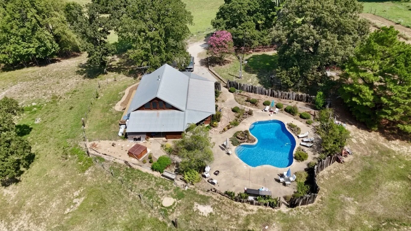 Curved swimming pool with concrete decking, surrounded by a wooden fence, mature trees, and a metal-roofed structure