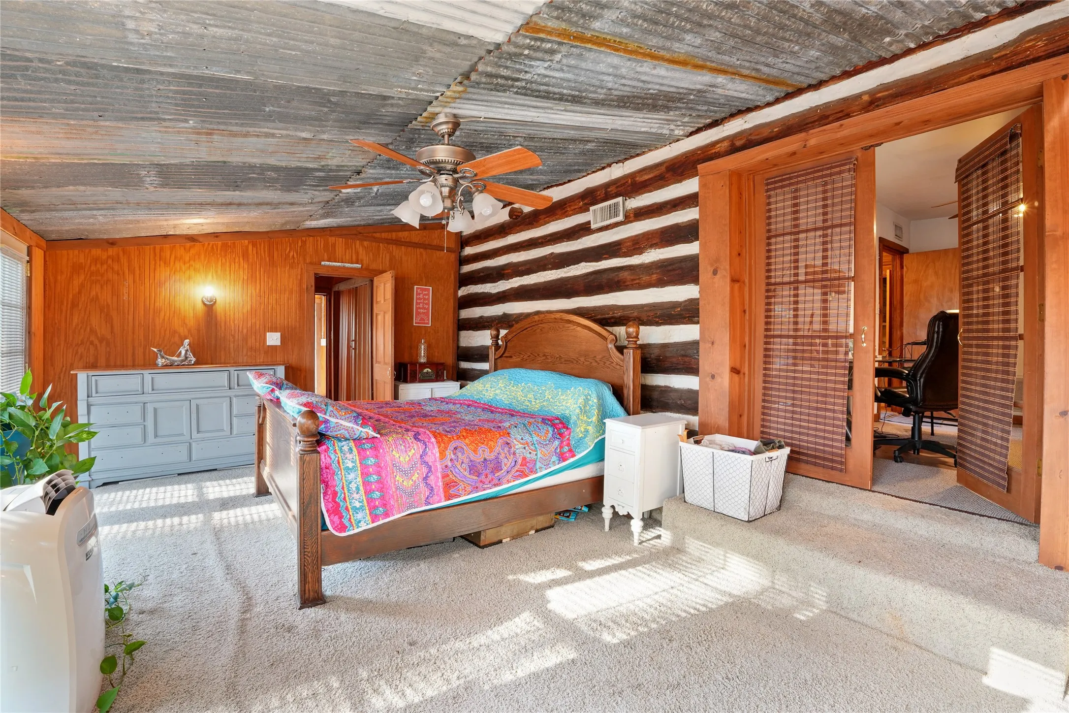Corrugated metal ceiling with a ceiling fan, log wall accent, wood-paneled walls, and light-toned carpet flooring