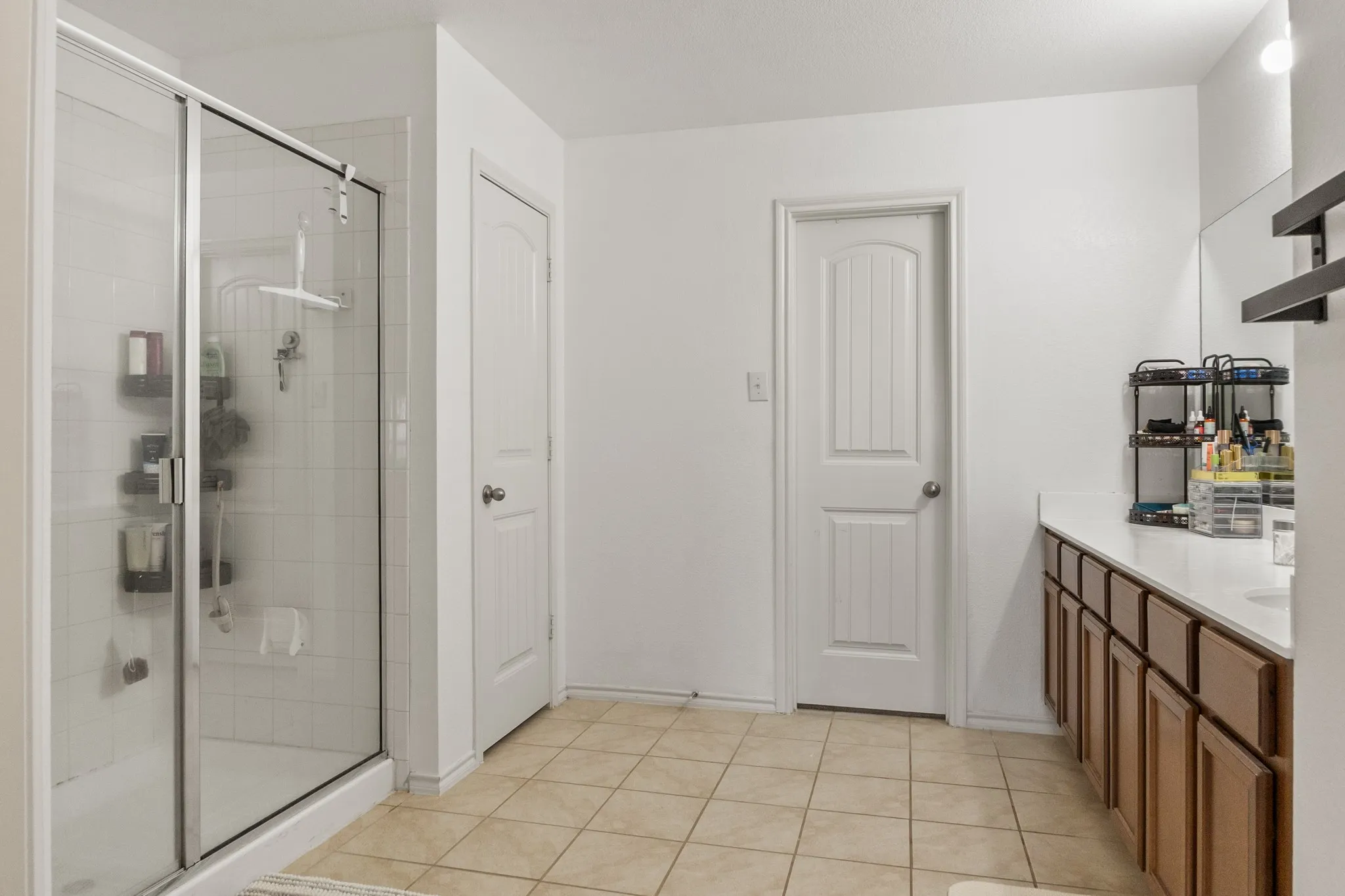 Tiled shower with glass enclosure, dual-sink vanity with white countertop, brown wood cabinetry, and neutral tile flooring