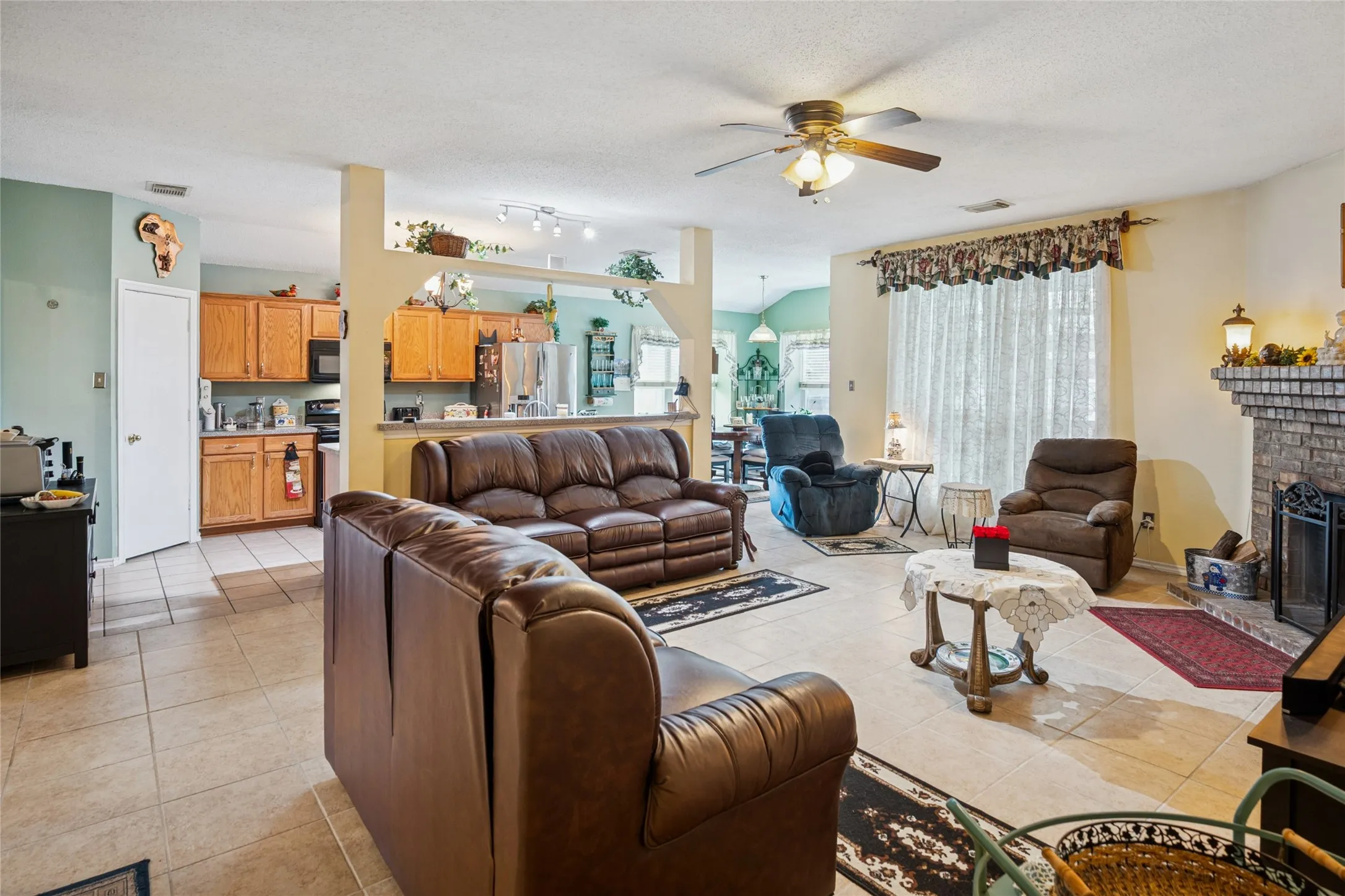Tiled flooring throughout, ceiling fan with integrated lighting, brick-faced fireplace open to kitchen with wood cabinetry.