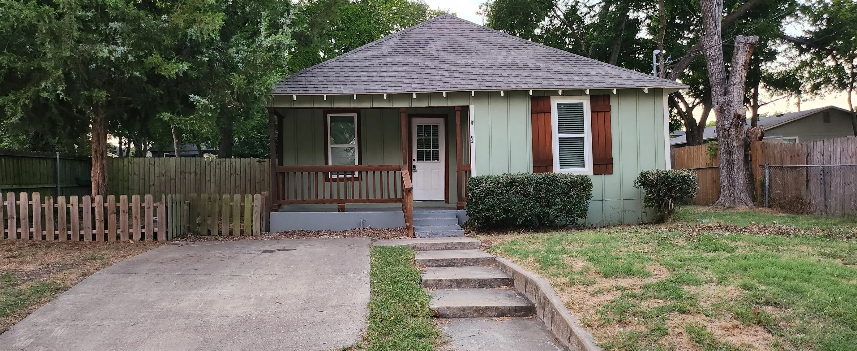Gabled roofline, light green siding, wood-toned shutters, front porch with wood railings, and a concrete pathway with steps