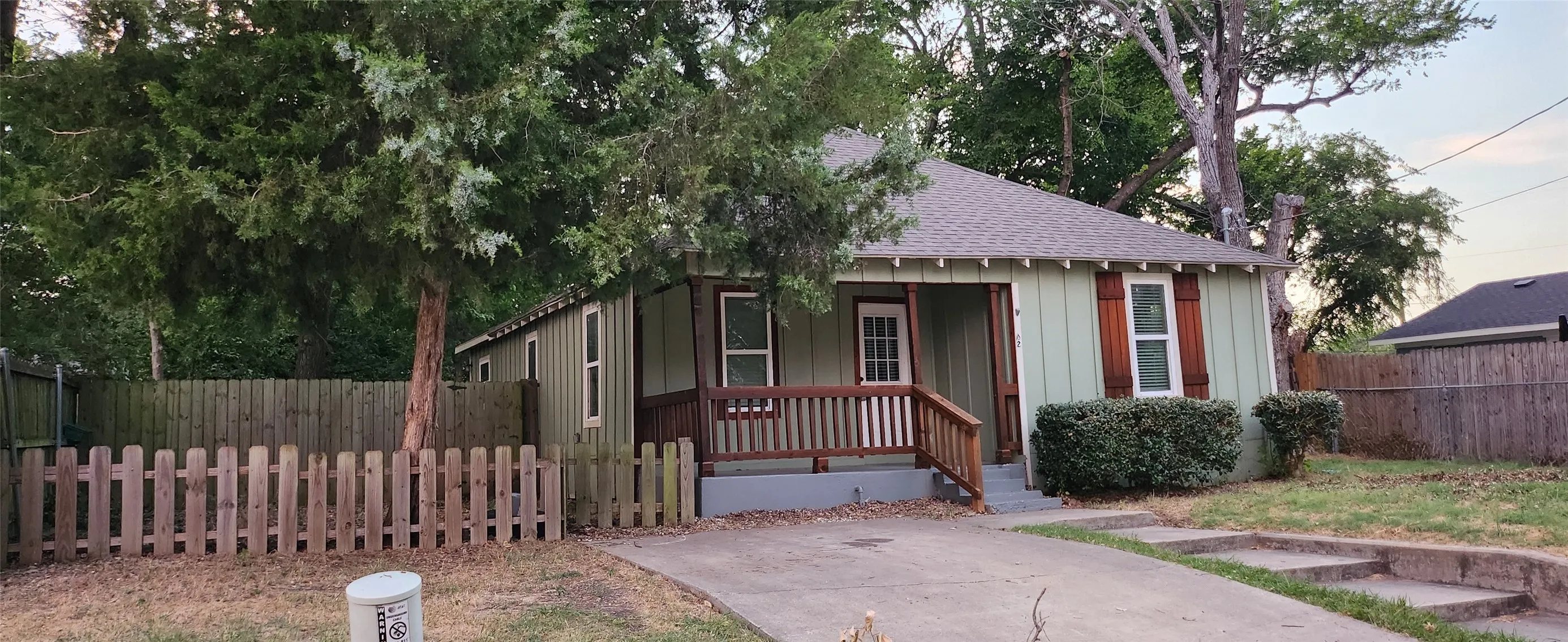 Light green siding exterior, covered porch with wood railing, wood shutters, fenced perimeter, and a concrete driveway