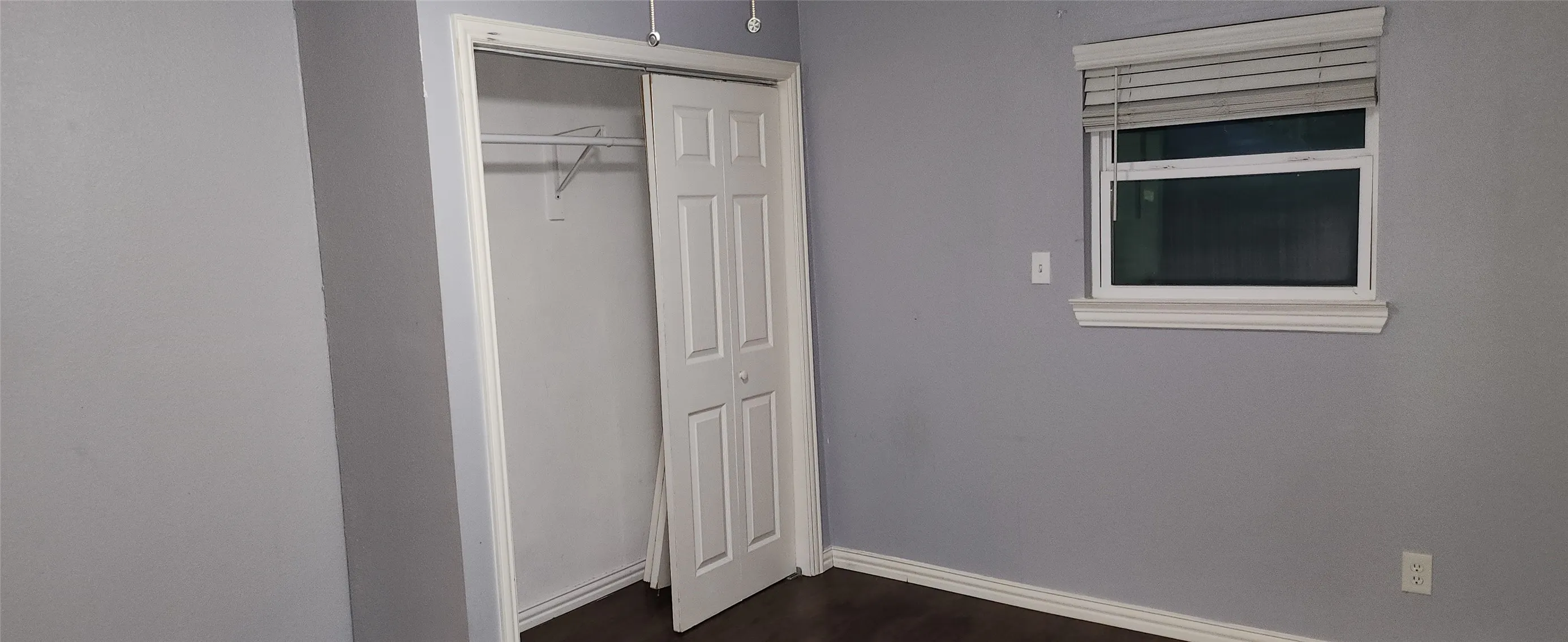 Neutral gray walls with wood-finish flooring, featuring a window with horizontal blinds and a bi-fold closet door