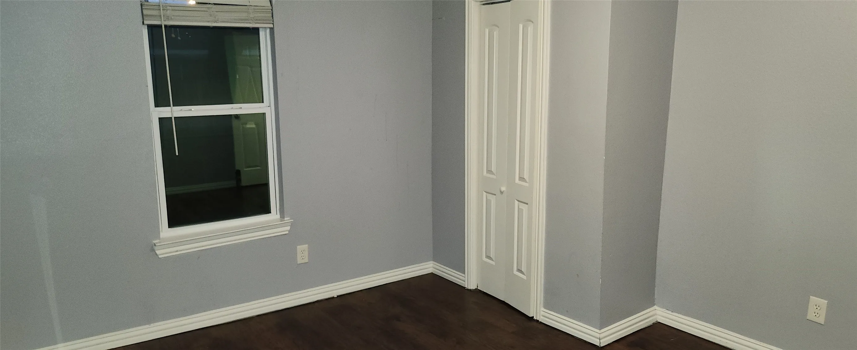 Grey painted walls, dark wood-finish flooring, white window trim, white bi-fold closet door, and white floor trim