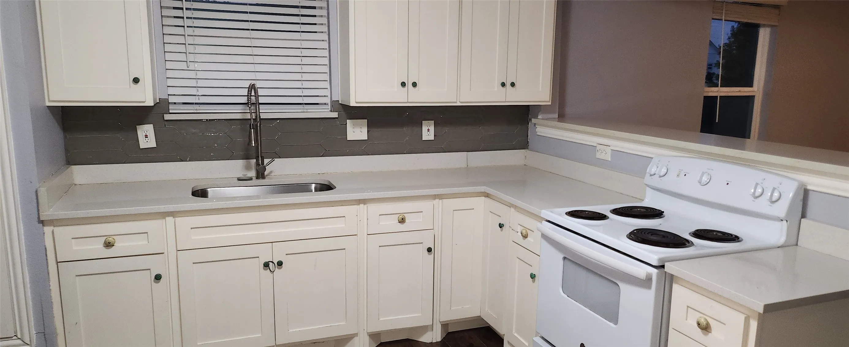 White cabinetry with dark hardware, a stainless steel sink with a high-arch faucet, a textured gray backsplash, light-toned countertops, and a white electric range