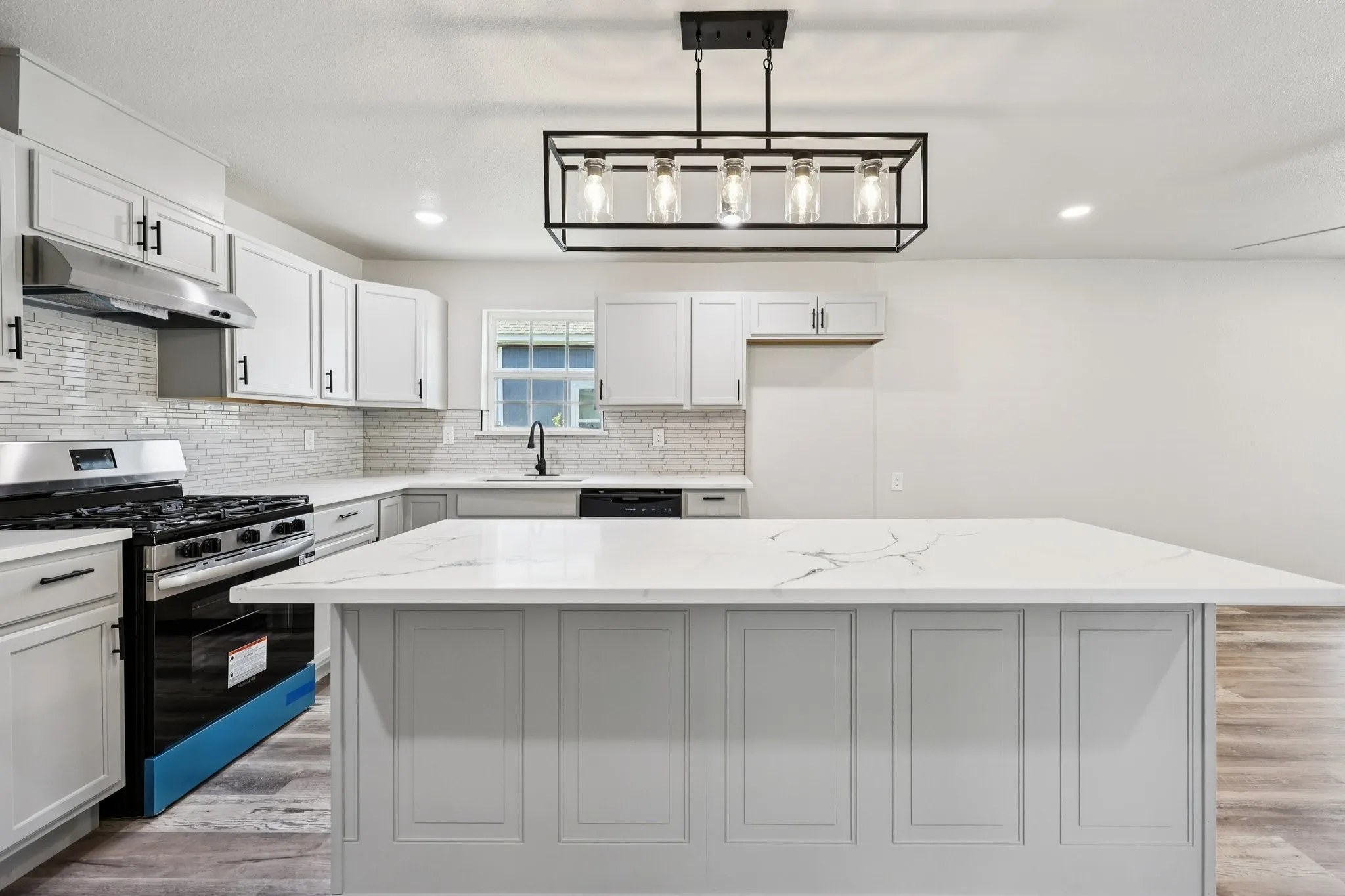Open-concept kitchen featuring an expansive island with a stone-finish countertop, light gray cabinetry, stainless steel appliances, a subway tile backsplash, and wood-finish flooring