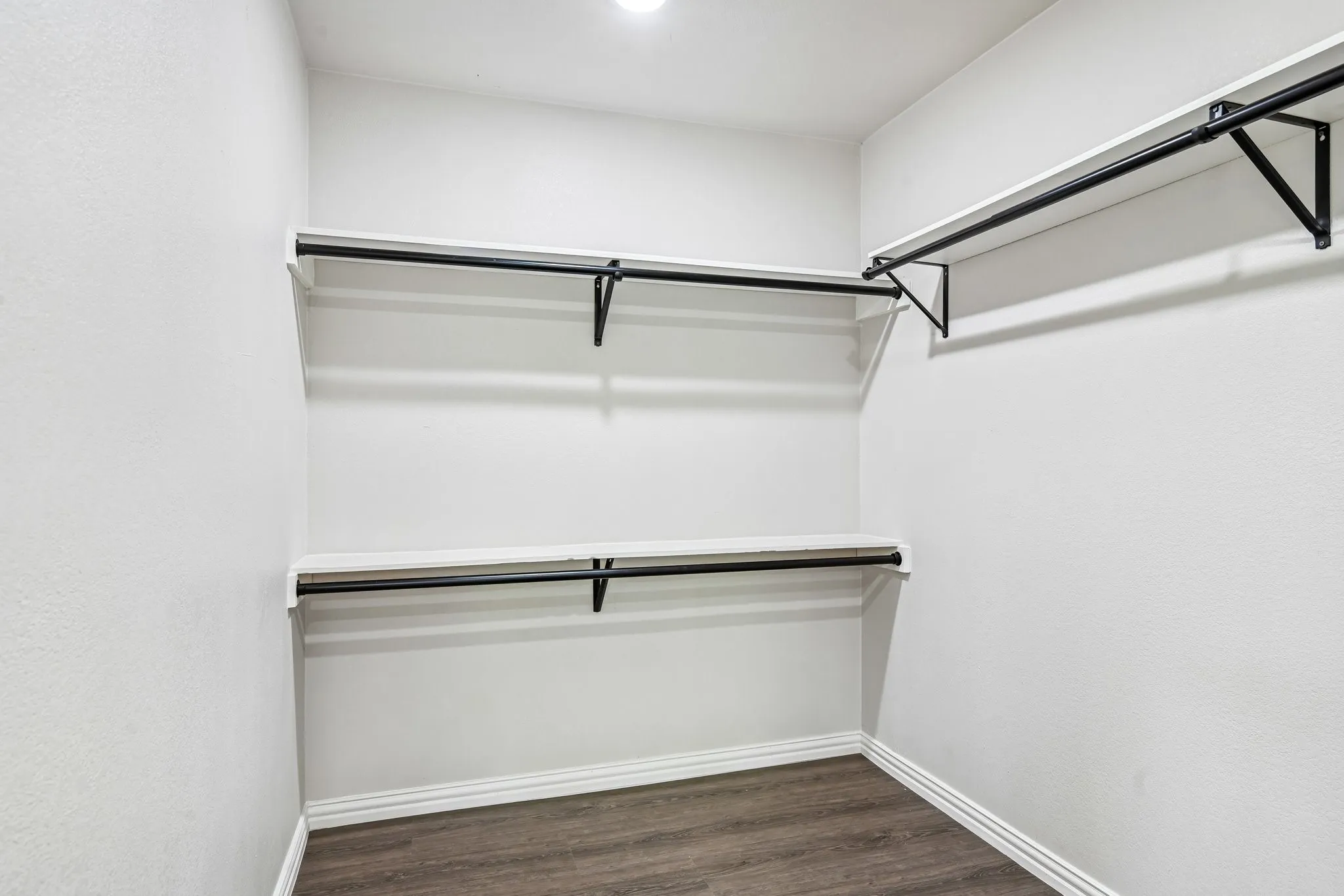 Walk-in closet featuring white walls, wood-finish flooring, white baseboards, two levels of shelving, and black metal hanging rods