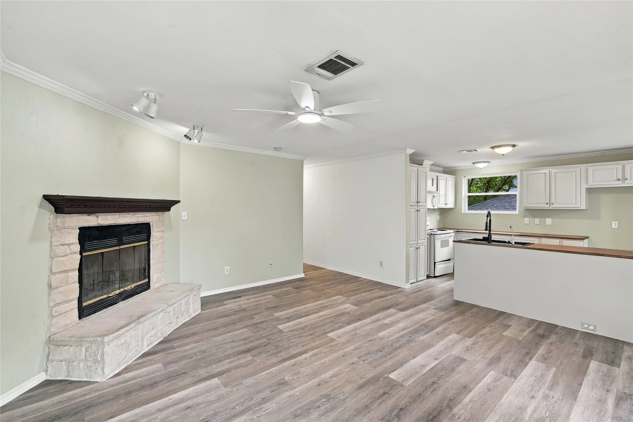 Stone-wrapped fireplace with a dark wood mantel, wood-look flooring, white kitchen cabinetry, and a ceiling fan with integrated lighting