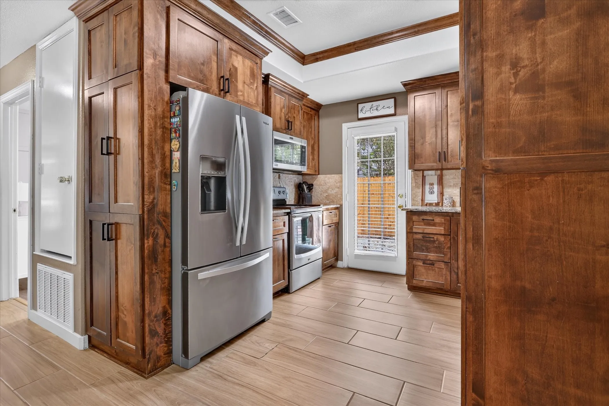 Wood-finish cabinetry, stainless steel appliances, tiled backsplash, light-toned flooring, and a glass-paned exterior door